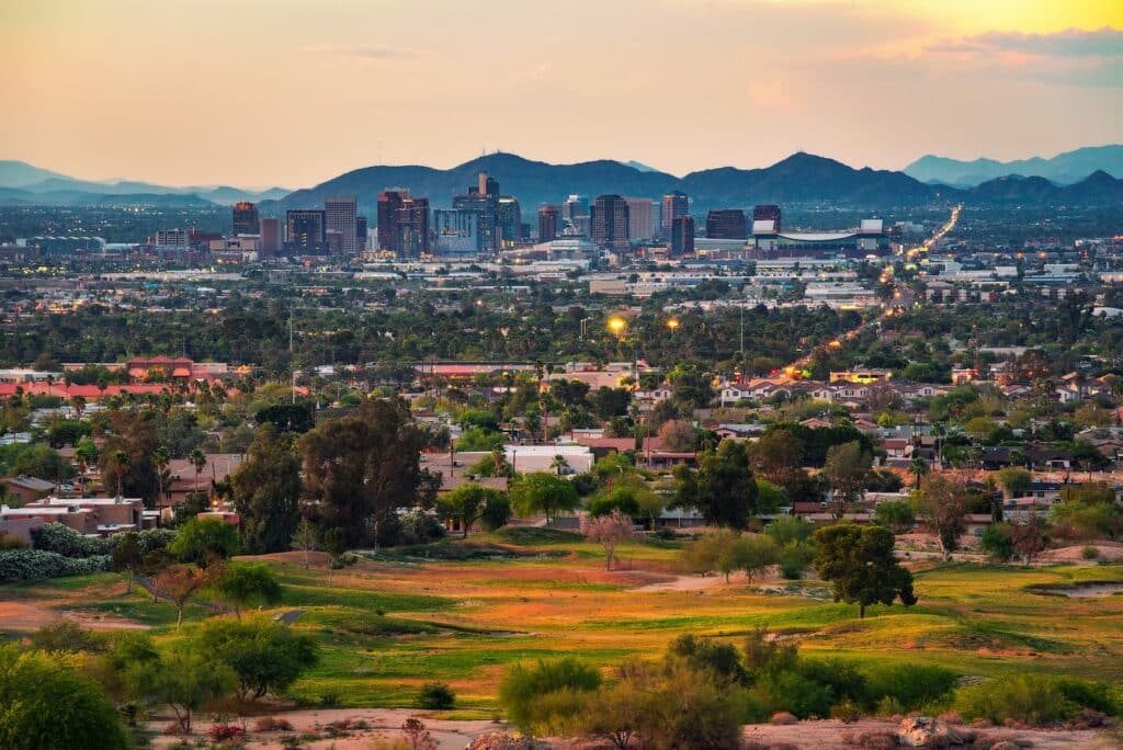 A city skyline with tall buildings is seen in the distance, surrounded by mountains, residential areas like Home - Scottsdale Entrada, and green spaces under a setting sun.