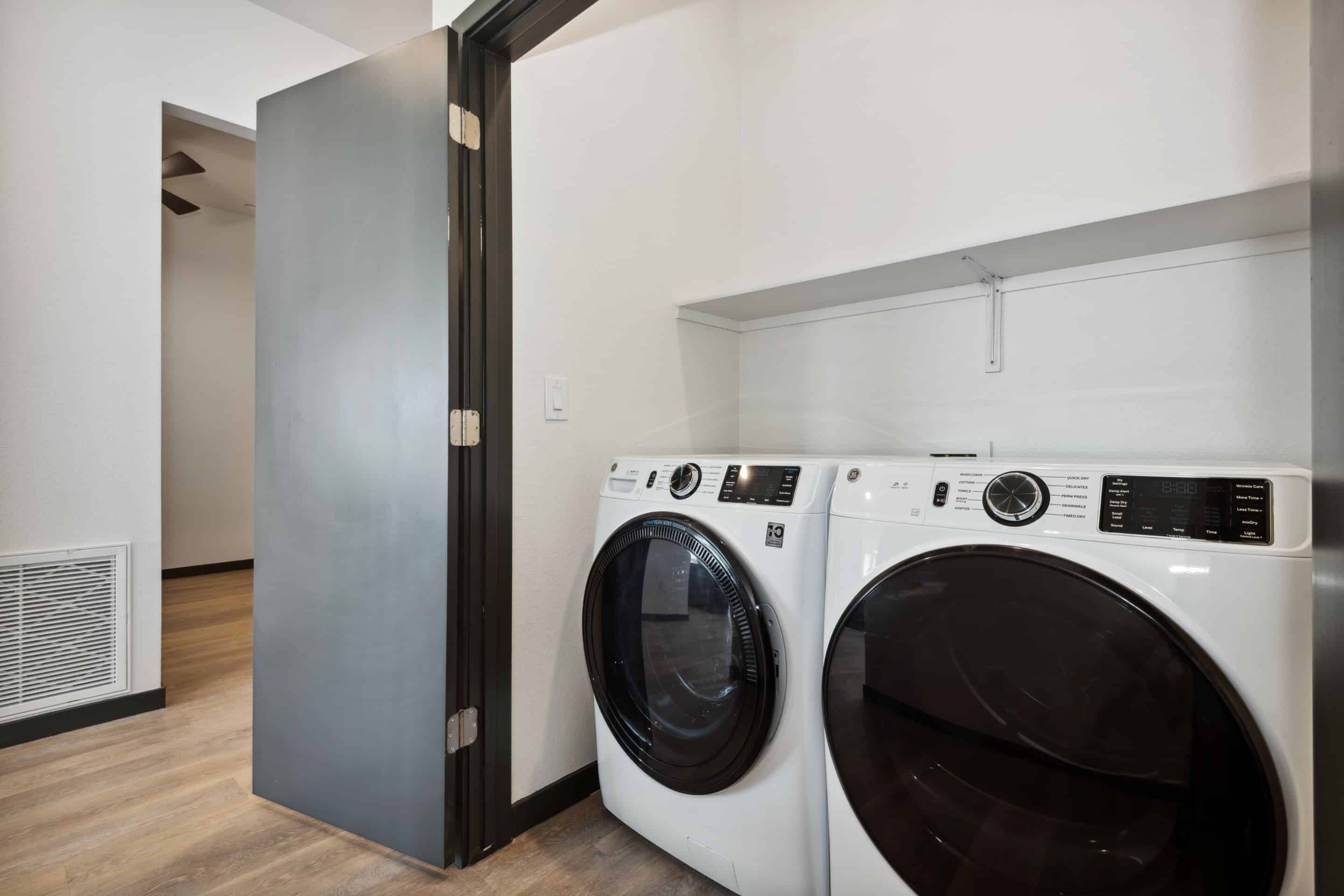 A modern laundry area at Home - Lake Cameron features a front-loading washer and dryer, white walls, wood flooring, and an open door leading to another room.
