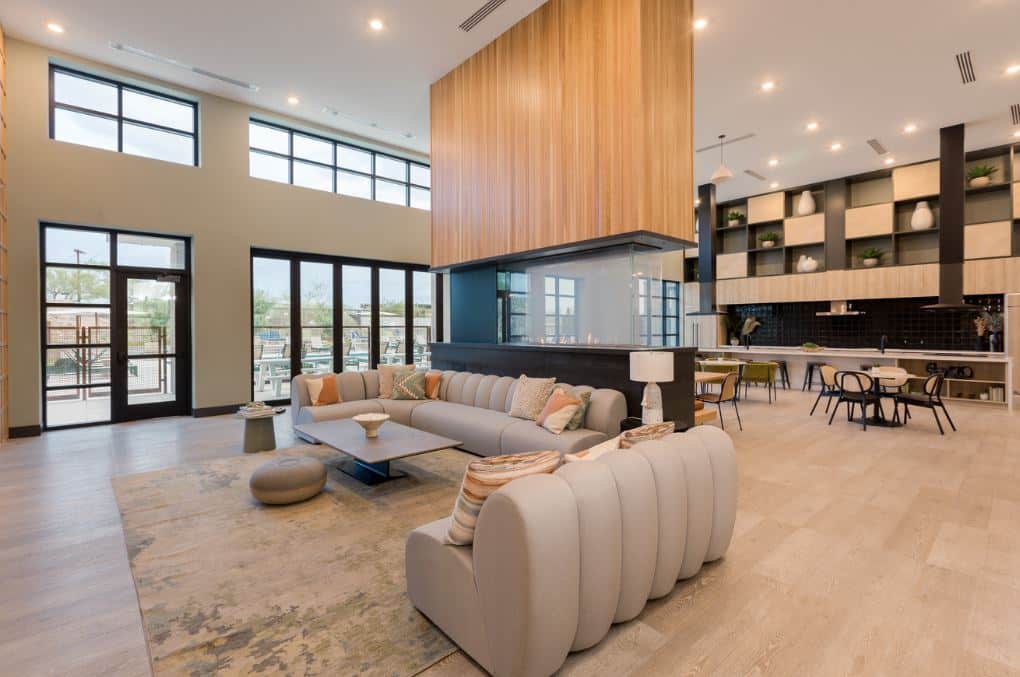 Modern open-concept lounge at Home - Lake Cameron, featuring a sectional sofa, large area rug, wood accents, floor-to-ceiling windows, and a kitchen with dining area in the background.