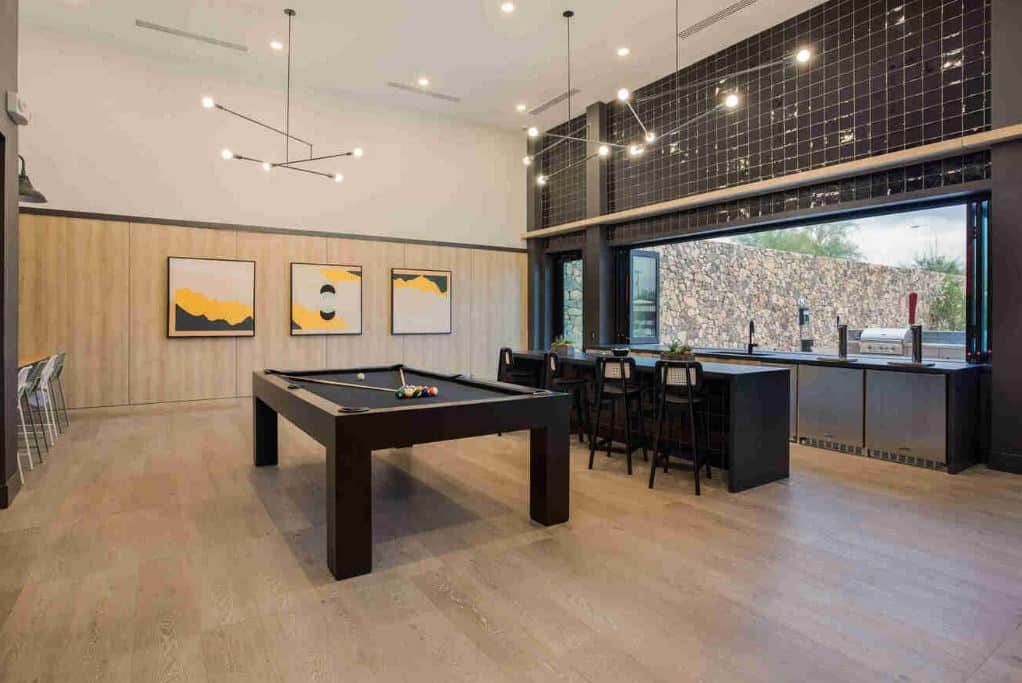 Modern game room at Home - Lake Cameron with a black pool table, bar seating, wall art, large windows, and views of the outdoor grill area through a sliding glass wall.