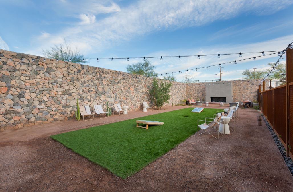 Outdoor courtyard at Home - Lake Cameron with artificial turf, string lights, seating, stone walls, a fireplace, and cornhole boards on the grass under a partly cloudy sky.