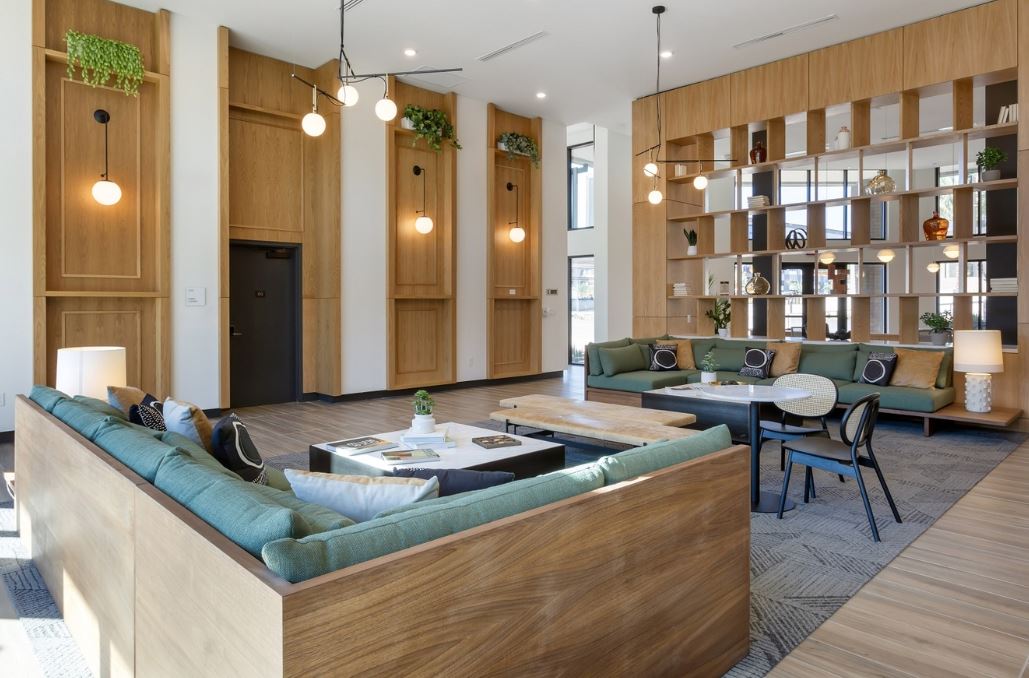 A modern lounge area at Home - Lake Cameron, featuring green sectional sofas, wooden paneling, bookshelves, round tables, and hanging light fixtures.