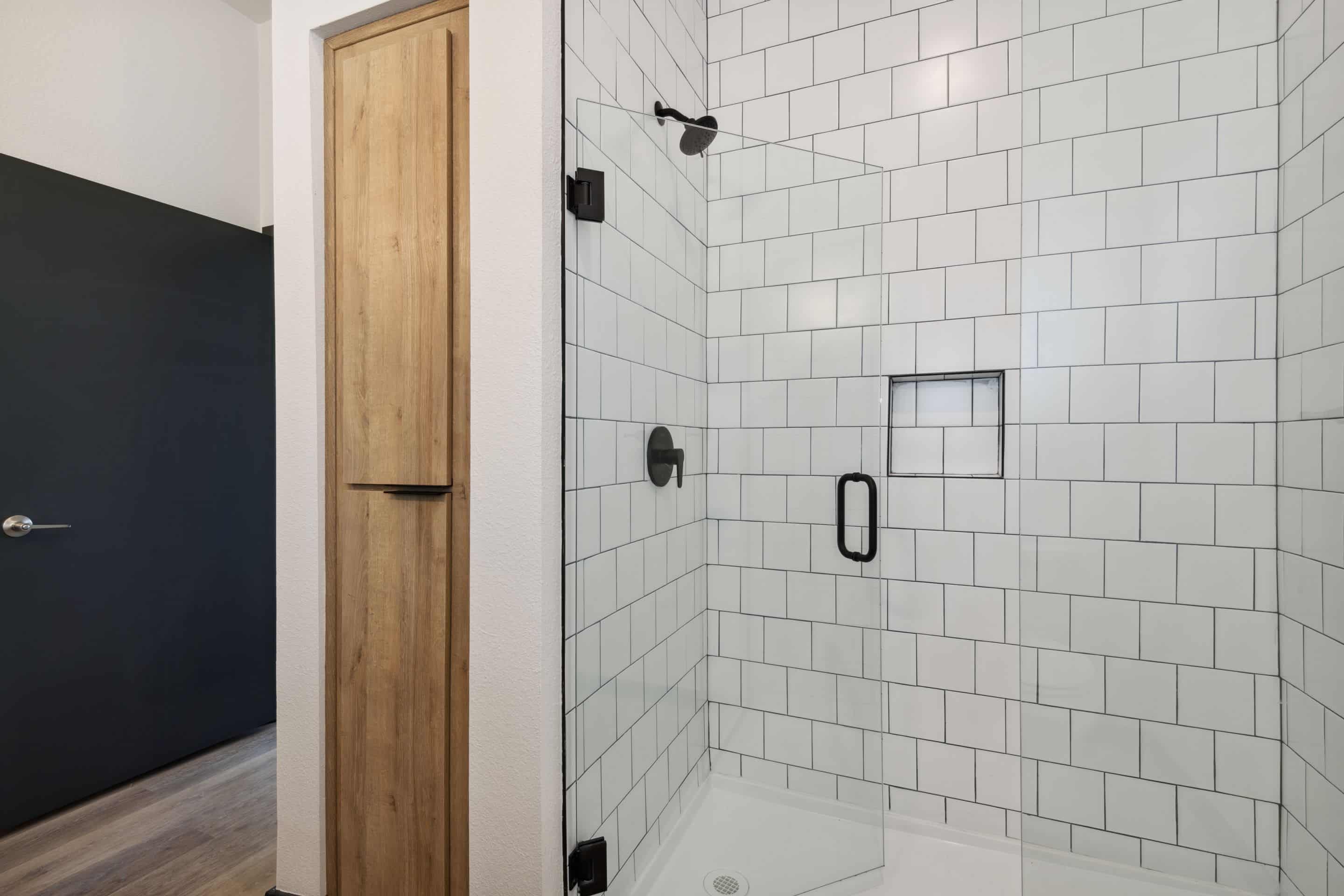 A modern bathroom shower at Home - Lake Cameron features white subway tile walls, black fixtures, a glass door, and a built-in wall niche. A wooden cabinet is visible beside the shower.