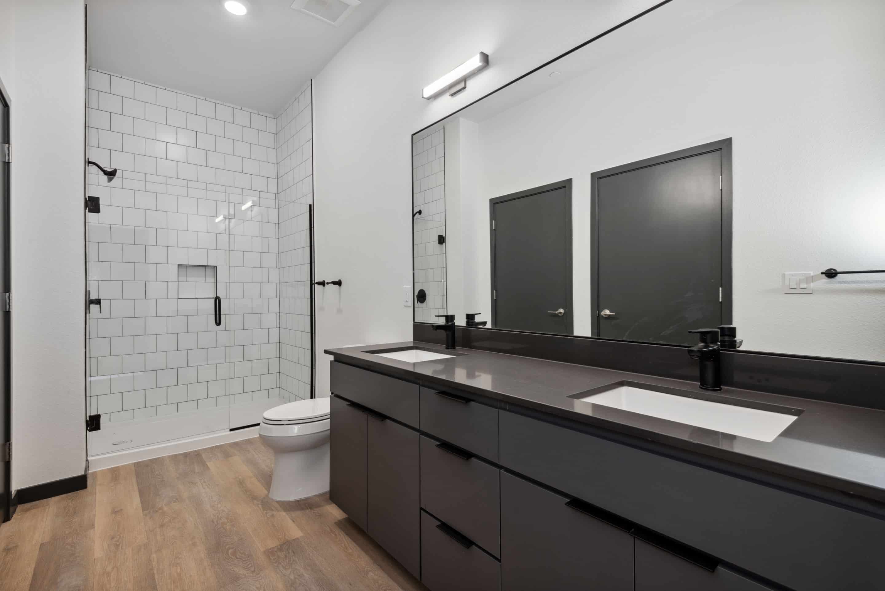 Modern bathroom at Home - Lake Cameron with a double-sink vanity, dark cabinets, large mirror, glass-enclosed shower with white subway tiles, and a toilet; light wood flooring and black fixtures.