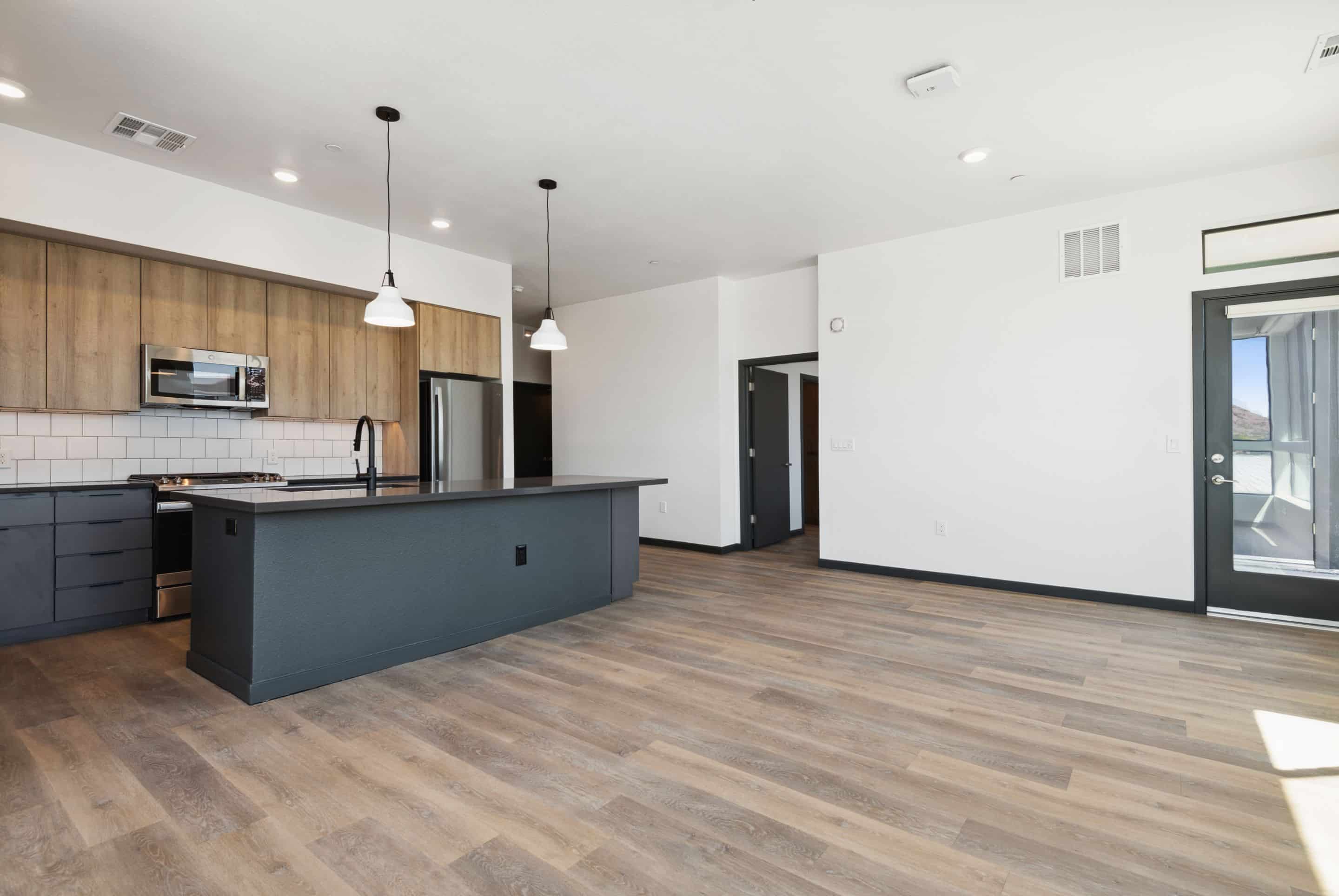 Modern open-concept kitchen and living area in this Home - Lake Cameron with wood flooring, black island, wood cabinets, stainless appliances, white tile backsplash, and pendant lights.