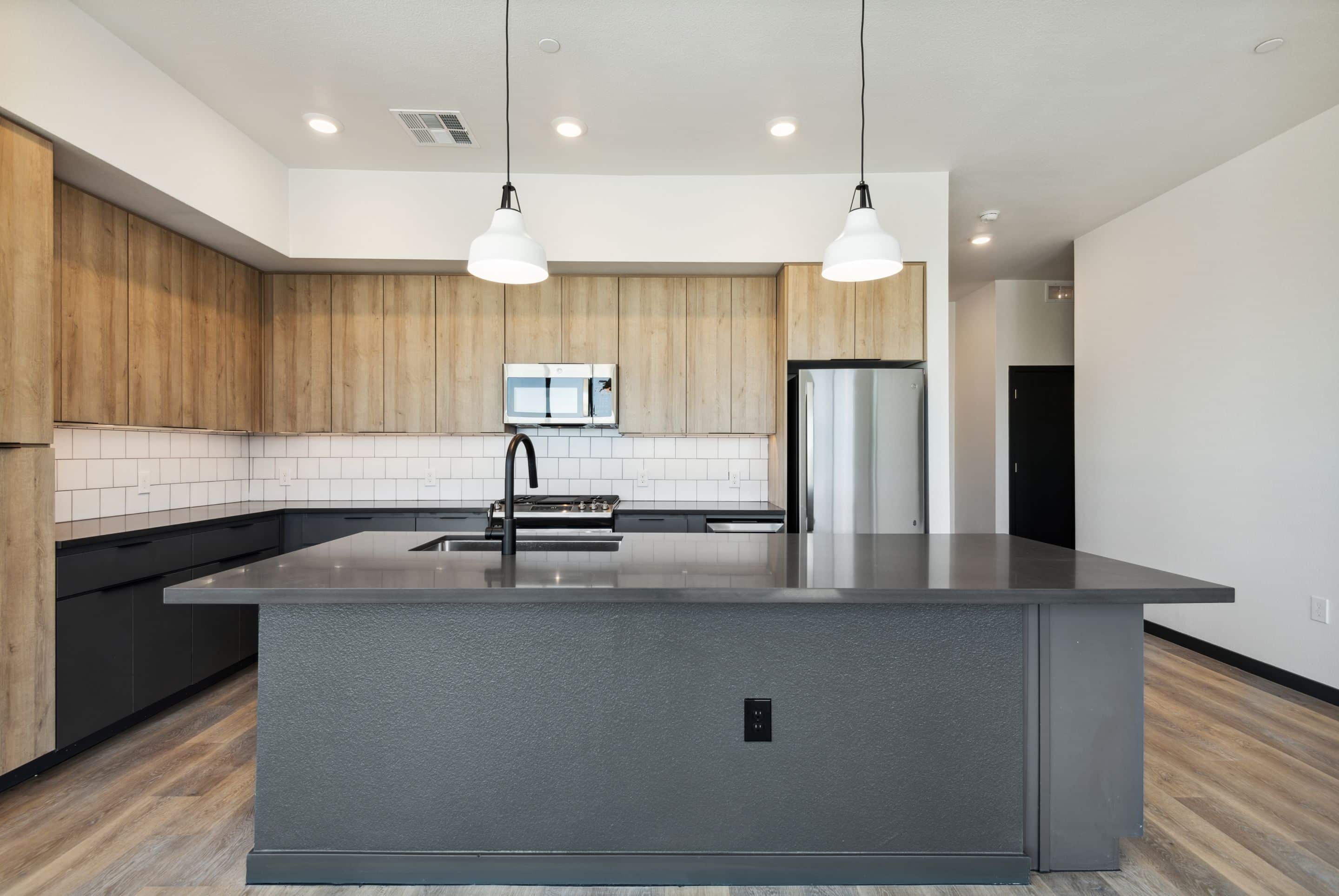 Modern kitchen in Home - Lake Cameron with wood cabinets, white tile backsplash, stainless steel appliances, large gray island, two pendant lights, and light wood flooring.