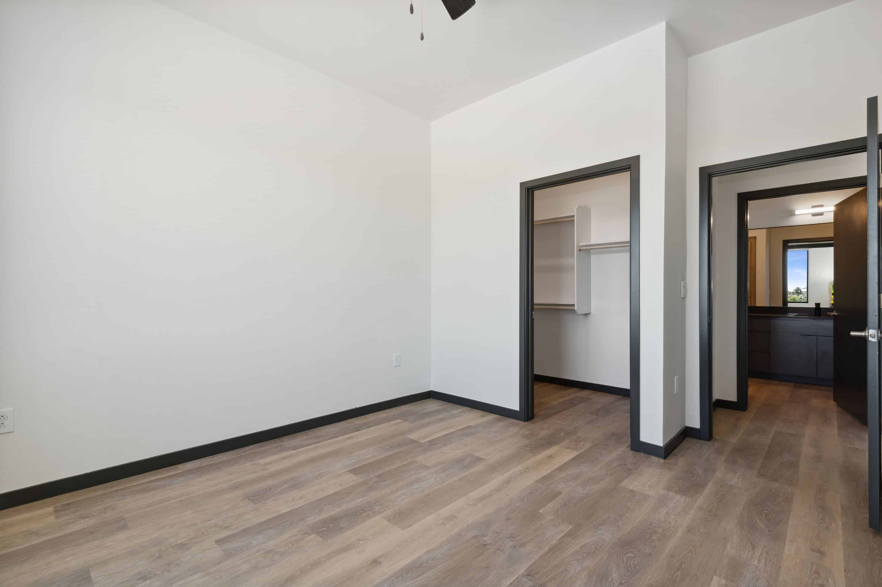 Empty room in a Home at Lake Cameron featuring light wood flooring, white walls with black trim, an open closet, and a doorway leading to another room with a visible window.