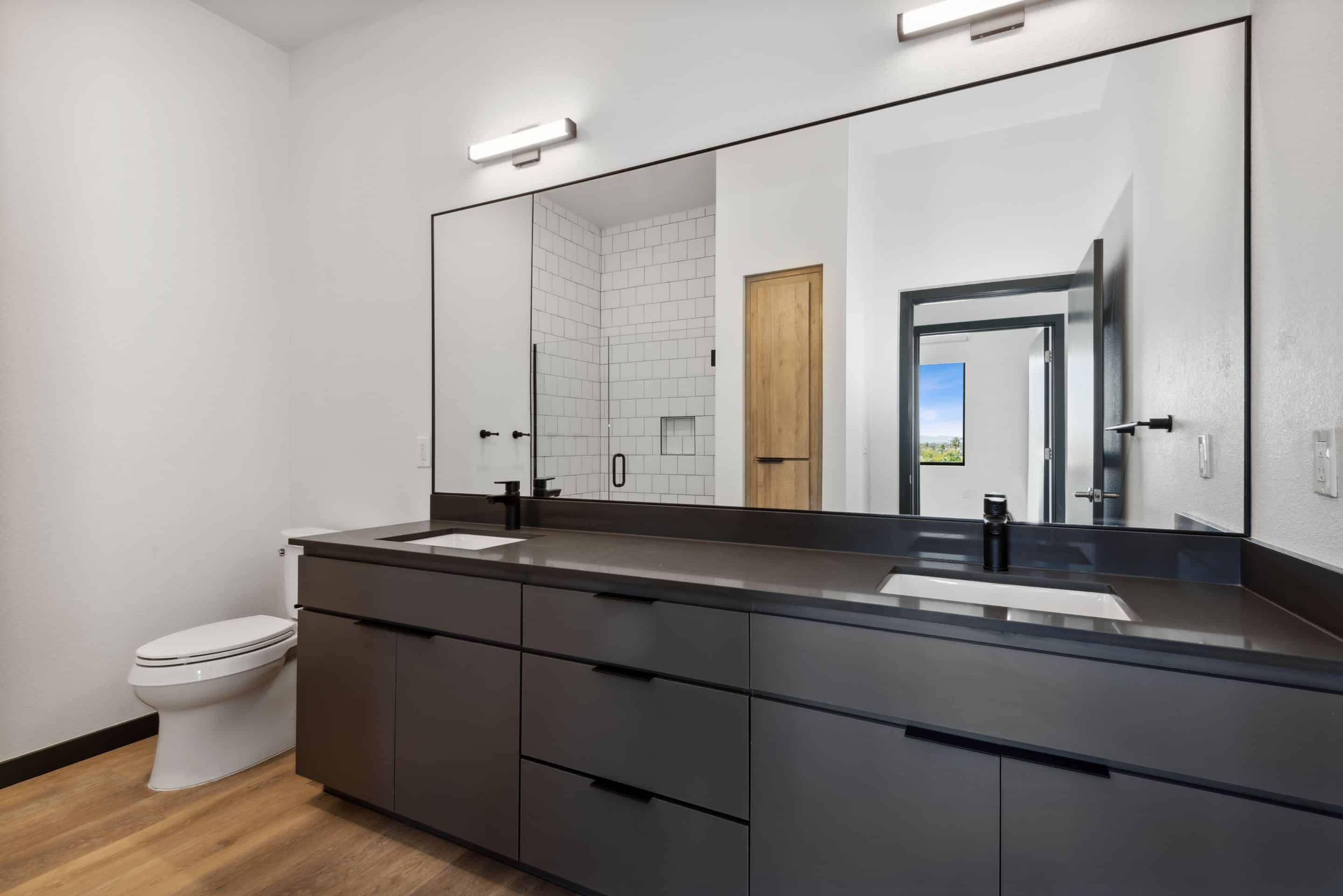 Modern bathroom in Home - Lake Cameron features a large mirror, double sinks, black countertop, wood flooring, and a white toilet. A walk-in shower with crisp white tiles is visible in the background.