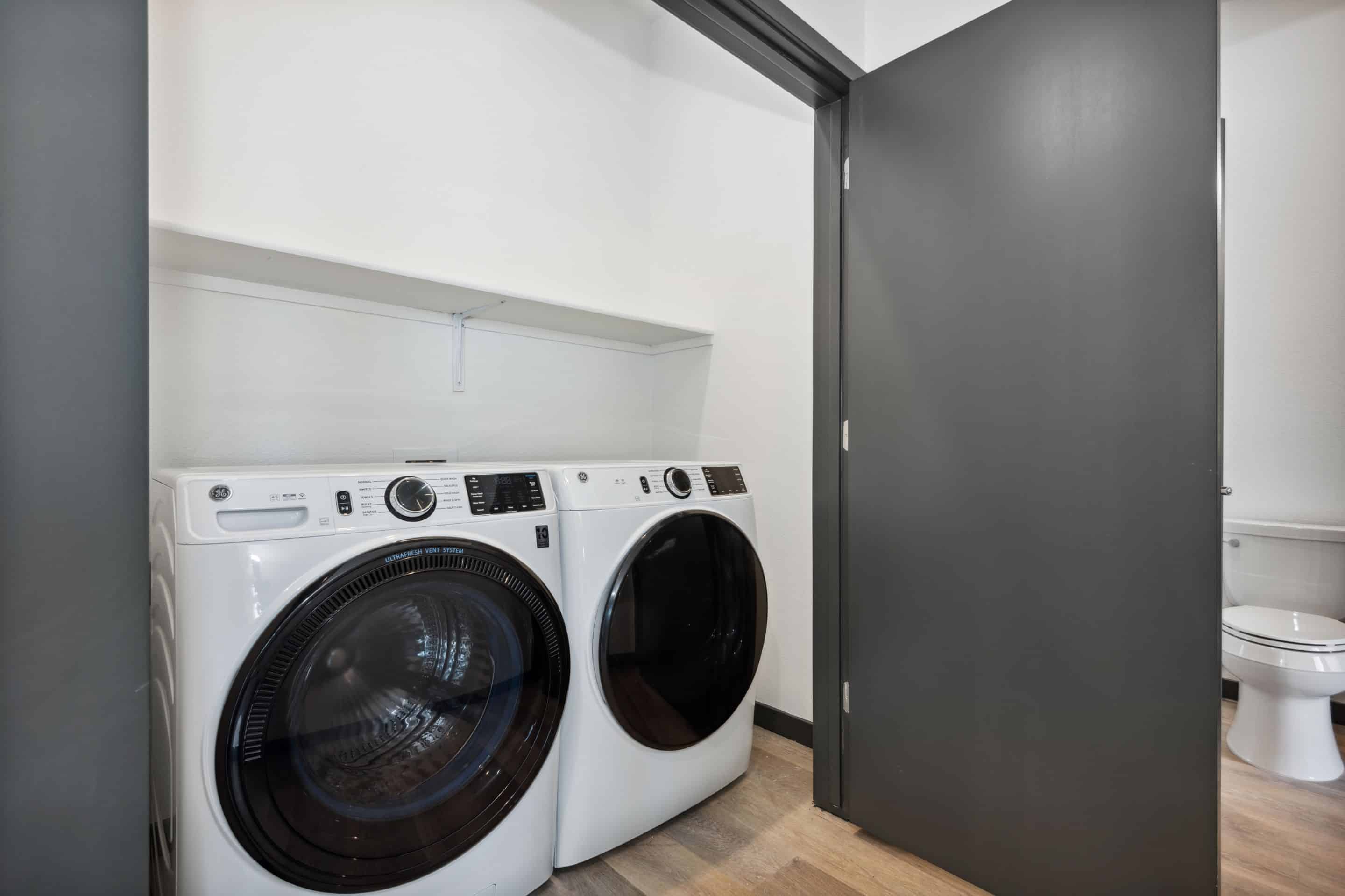 A laundry room in Home - Lake Cameron features a front-loading washer and dryer beneath a white shelf, next to an open door leading to a bathroom with a visible toilet.