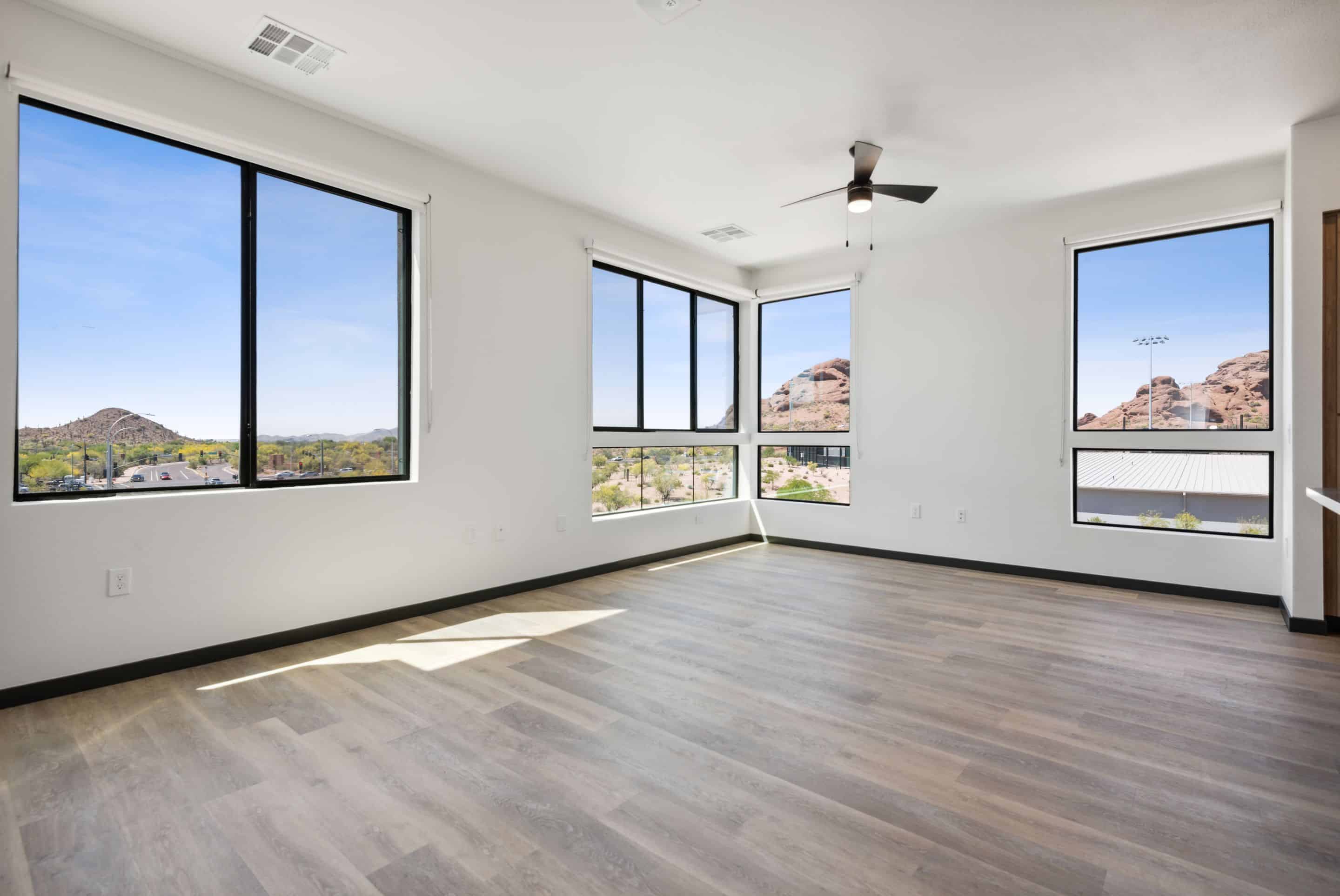 Empty modern room with large windows, light wood flooring, white walls, and ceiling fan; outside are rocky hills and a clear blue sky, offering scenic views perfect for a tranquil Home - Lake Cameron retreat.