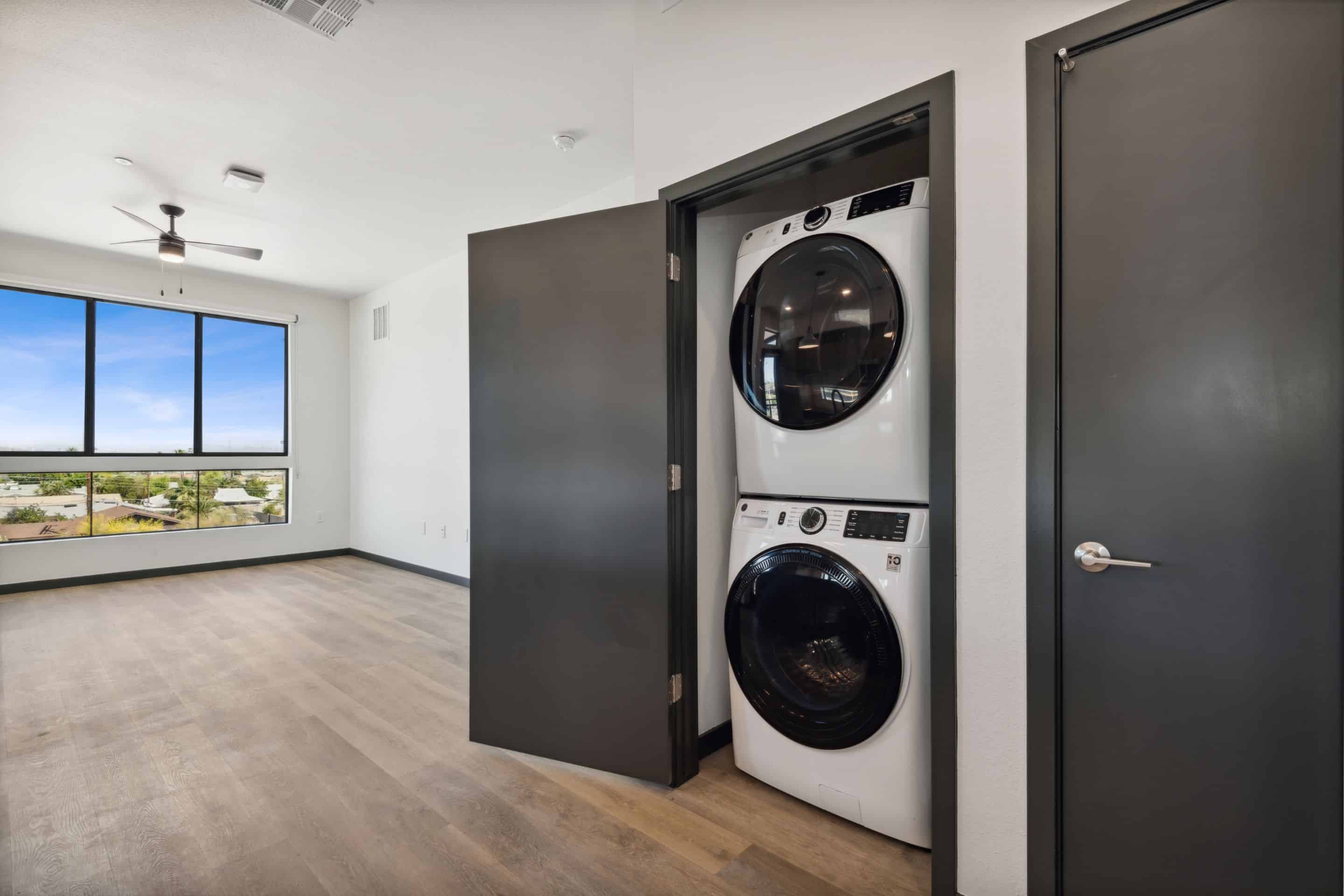 A modern apartment room at Home - Lake Cameron features wood floors, large windows, and an open closet with a stacked washer and dryer next to a closed dark door.
