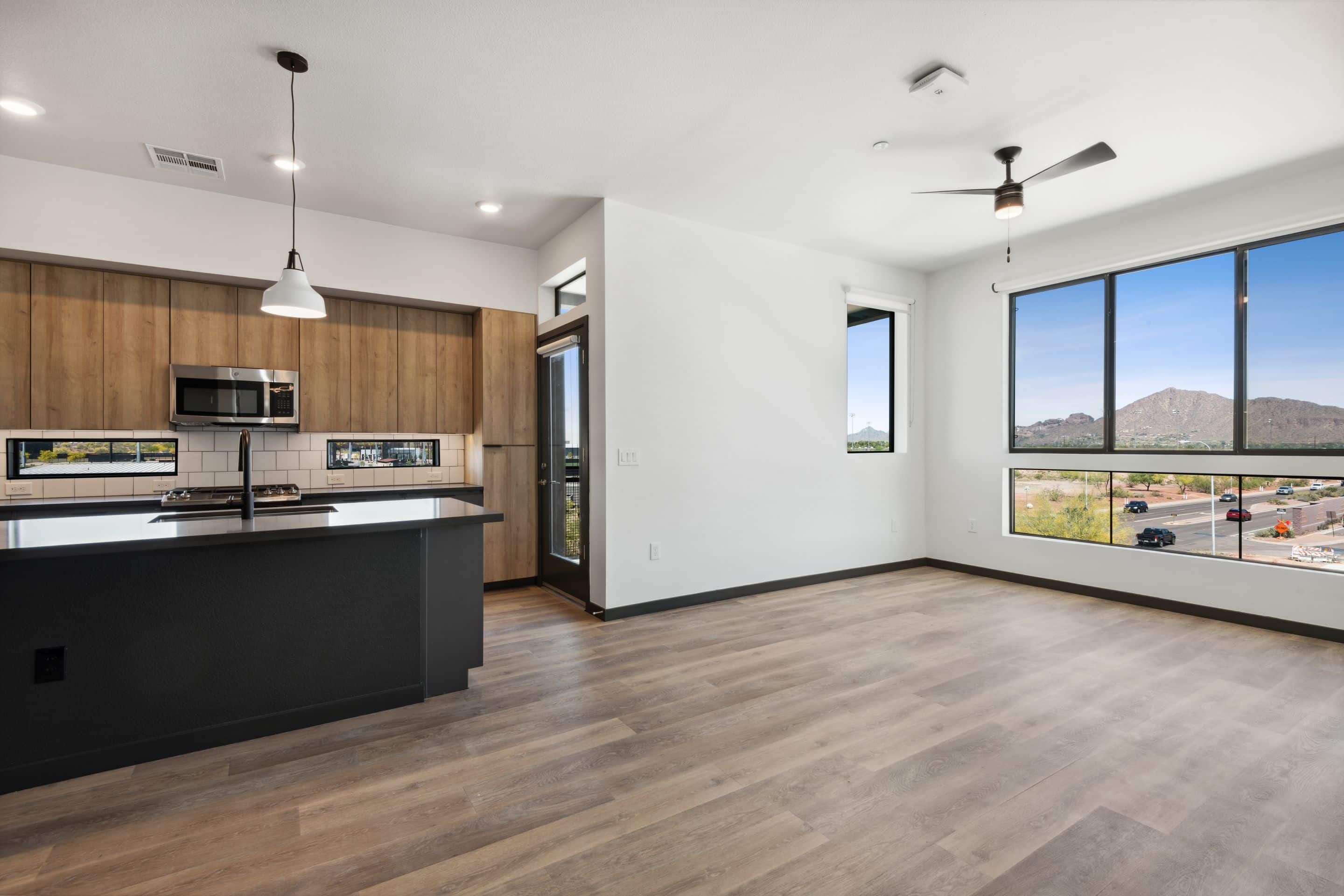 Modern open-concept kitchen and living area in Home - Lake Cameron, featuring wood floors, large windows with mountain views, and sleek black accents. Kitchen boasts wood cabinets, black countertops, and stainless steel appliances.