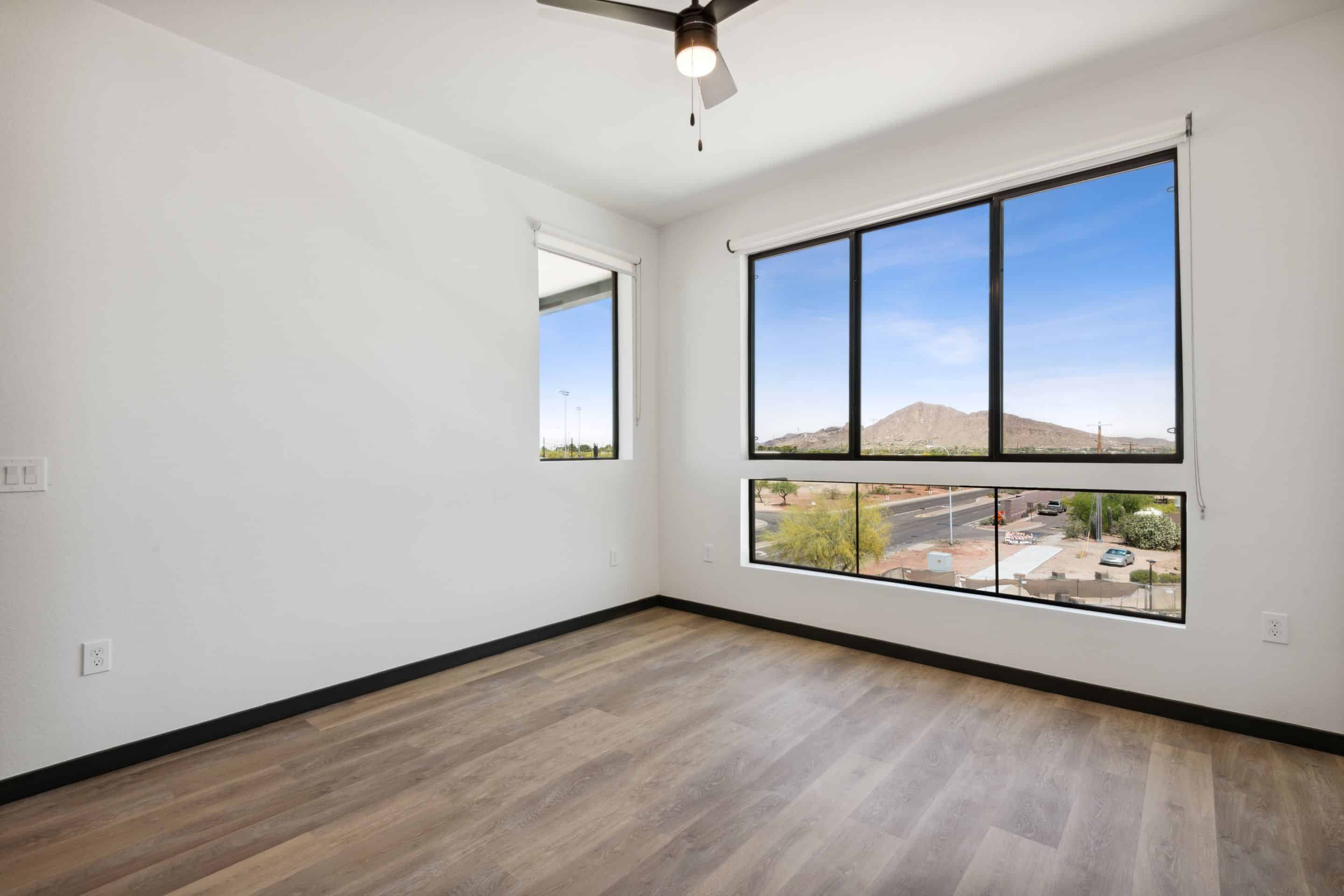 Bright and airy empty room with light wood flooring, white walls, a ceiling fan, and large windows offering views of parked cars, a road, and distant mountains—perfect for your new Home at Lake Cameron.