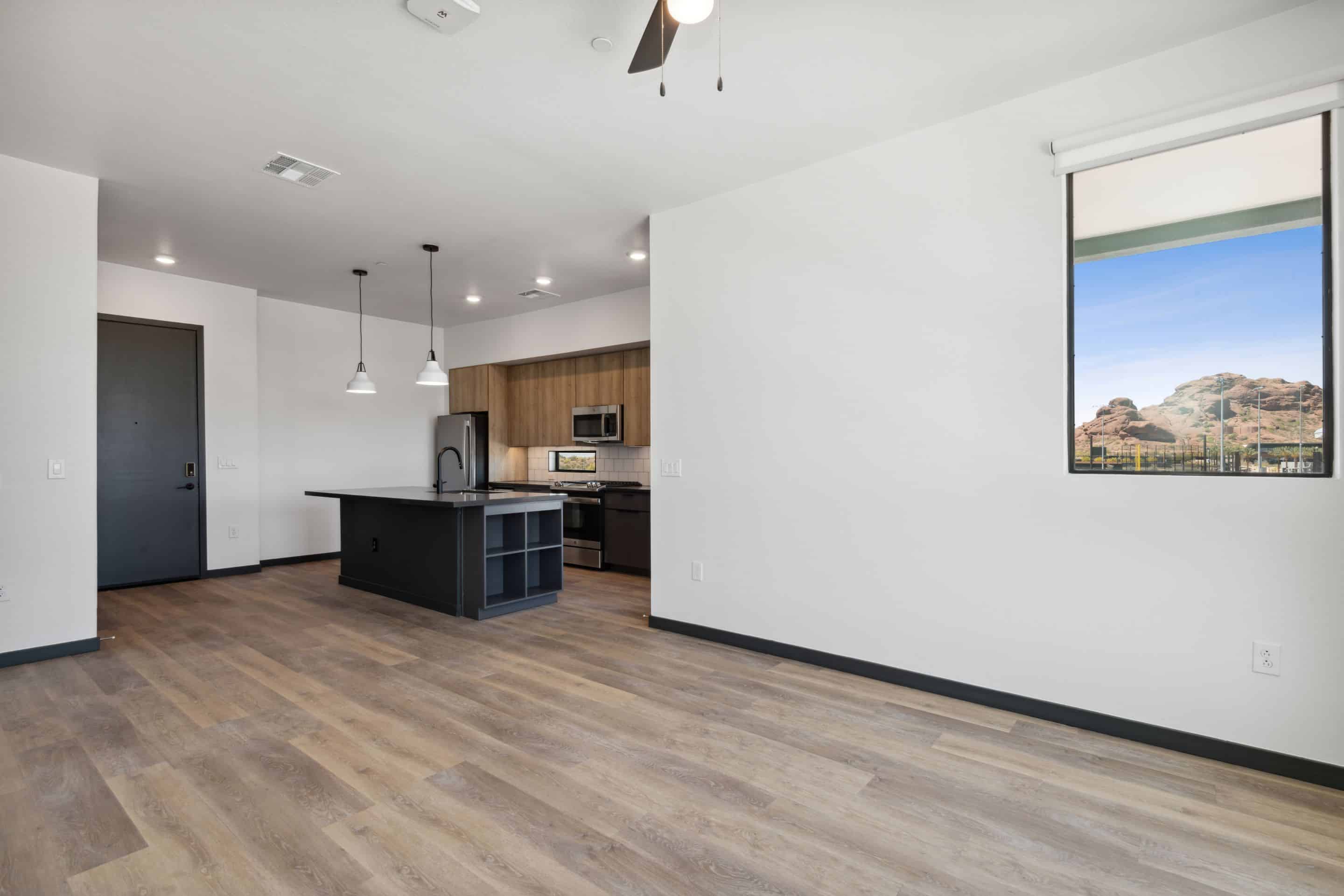 Modern empty apartment with light wood flooring, open kitchen with island, stainless steel appliances, and a window revealing a scenic rocky landscape—perfect for those seeking a tranquil home near Lake Cameron.