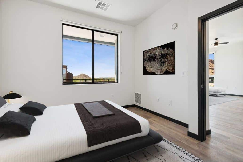 A modern bedroom at Home - Lake Cameron features a low bed, black and white bedding, abstract wall art, a window with a view, and an open door leading to another room.