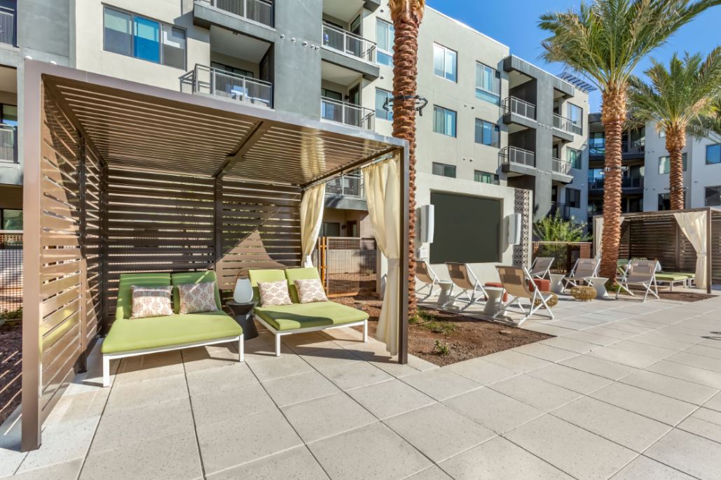 Outdoor lounge area at Home - Lake Cameron featuring cabanas, green cushioned chairs, palm trees, and apartment buildings in the background under a clear blue sky.