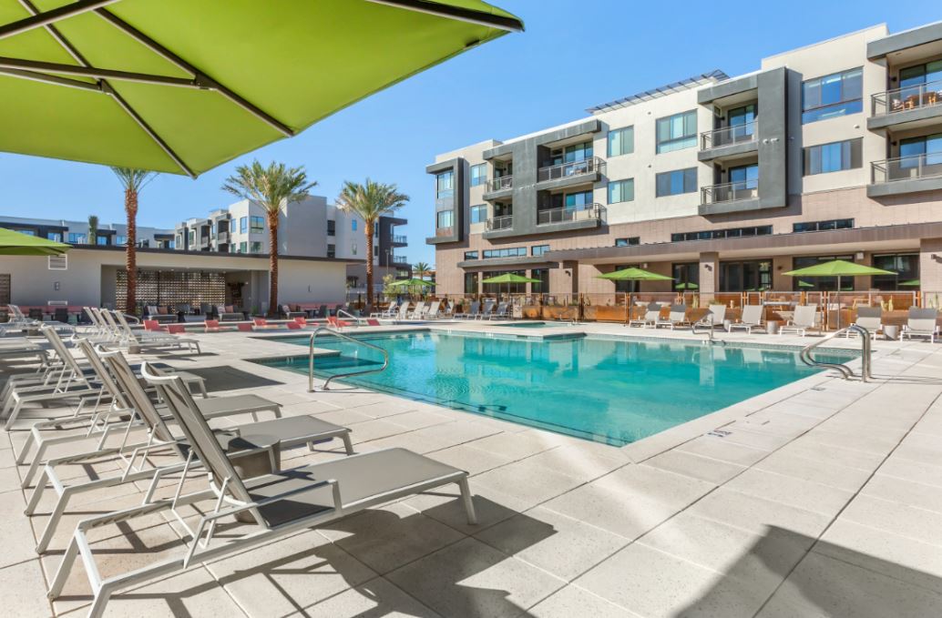 Modern apartment complex at Home - Lake Cameron features a central outdoor swimming pool, surrounded by lounge chairs, umbrellas, and palm trees under a clear blue sky.