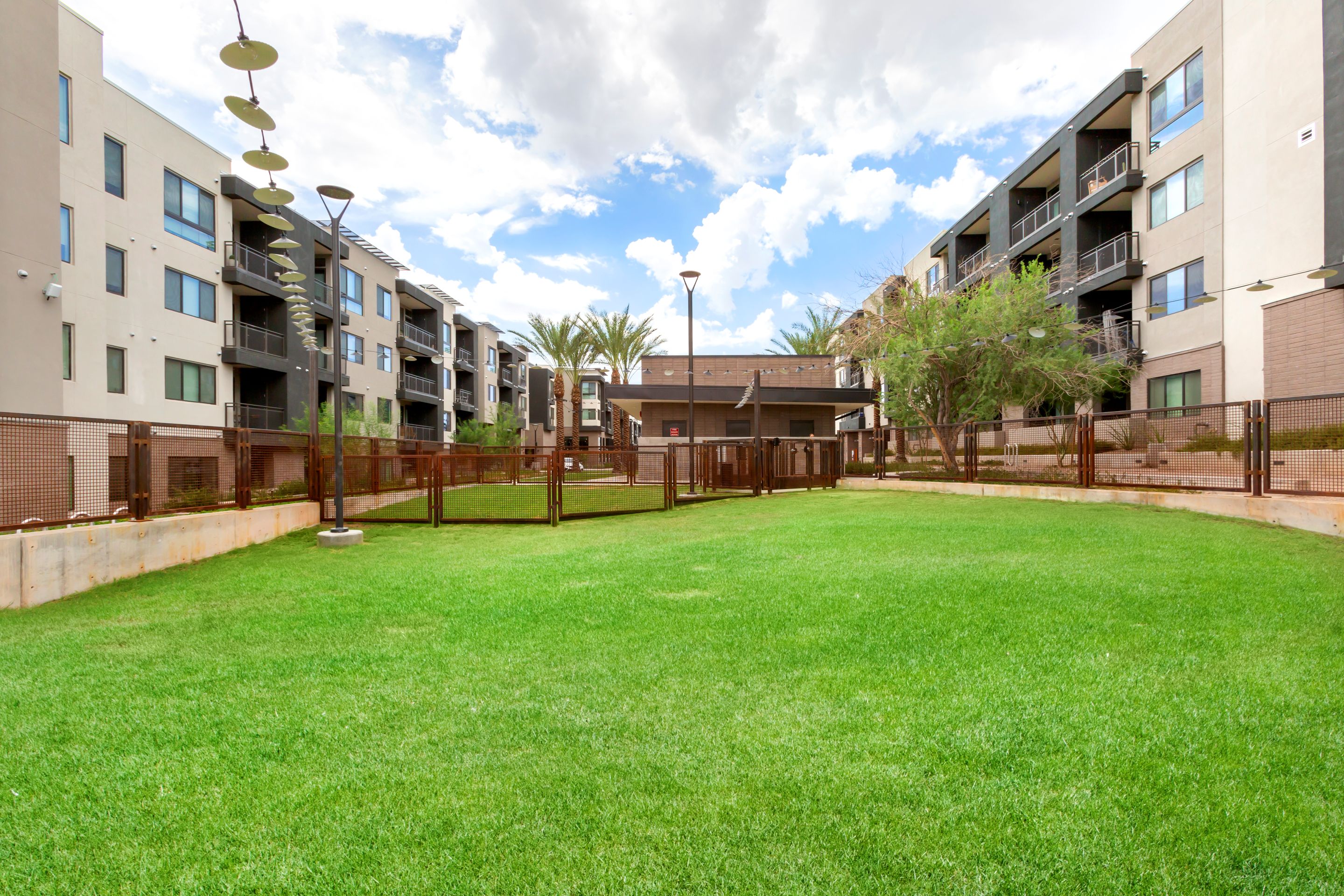 Fenced grassy area between modern apartment buildings at Home - Lake Cameron, with trees, a cloudy sky, and some outdoor lighting fixtures visible.