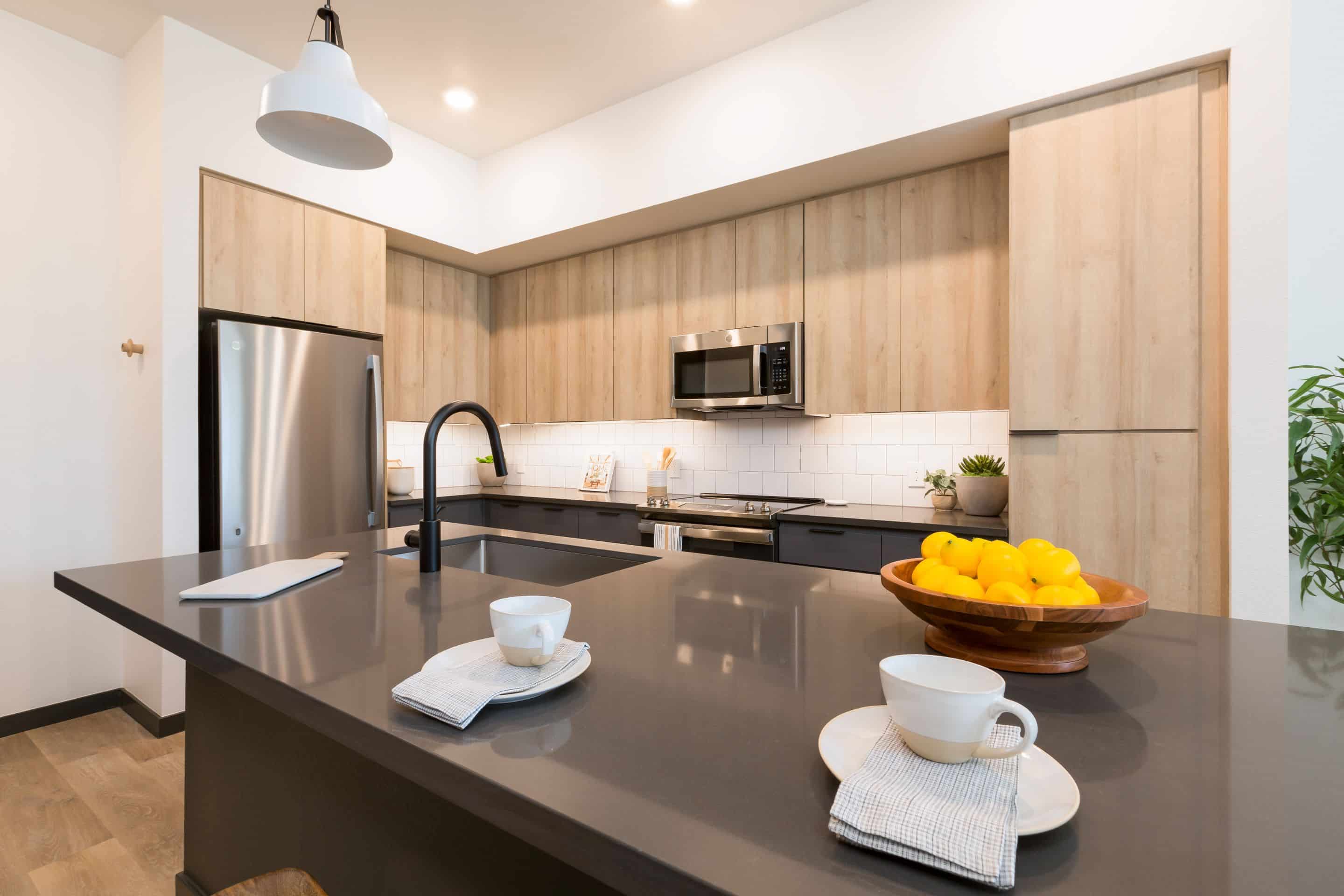 Modern kitchen with light wood cabinets, stainless steel appliances, black countertop, two cups on napkins, and a wooden bowl filled with yellow lemons on the island—perfect for a stylish home near Lake Cameron.
