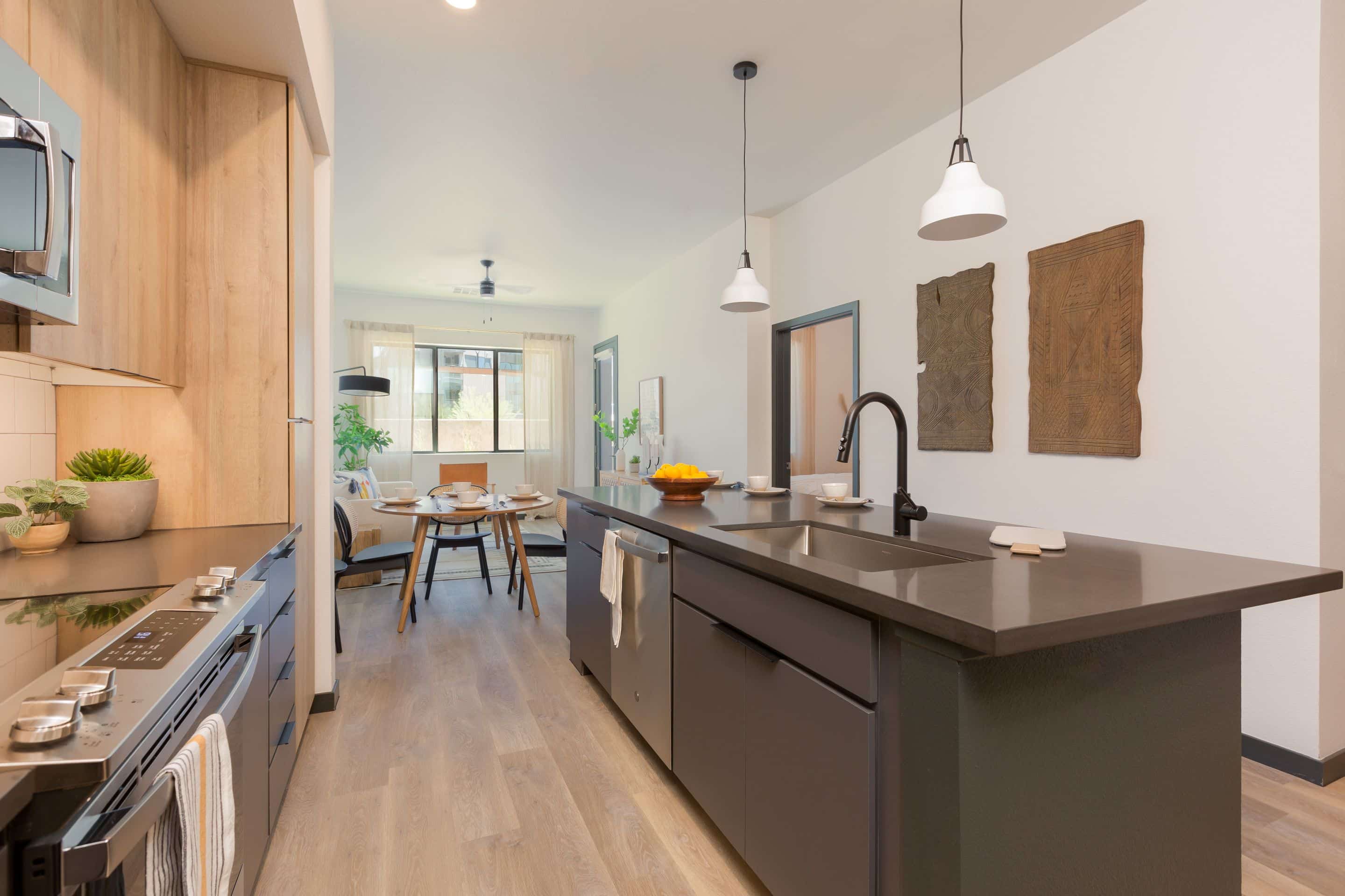 Modern kitchen in Home - Lake Cameron with a dark island, black faucet, wooden cabinets, and pendant lights, opening into a dining area with a round table and large window.
