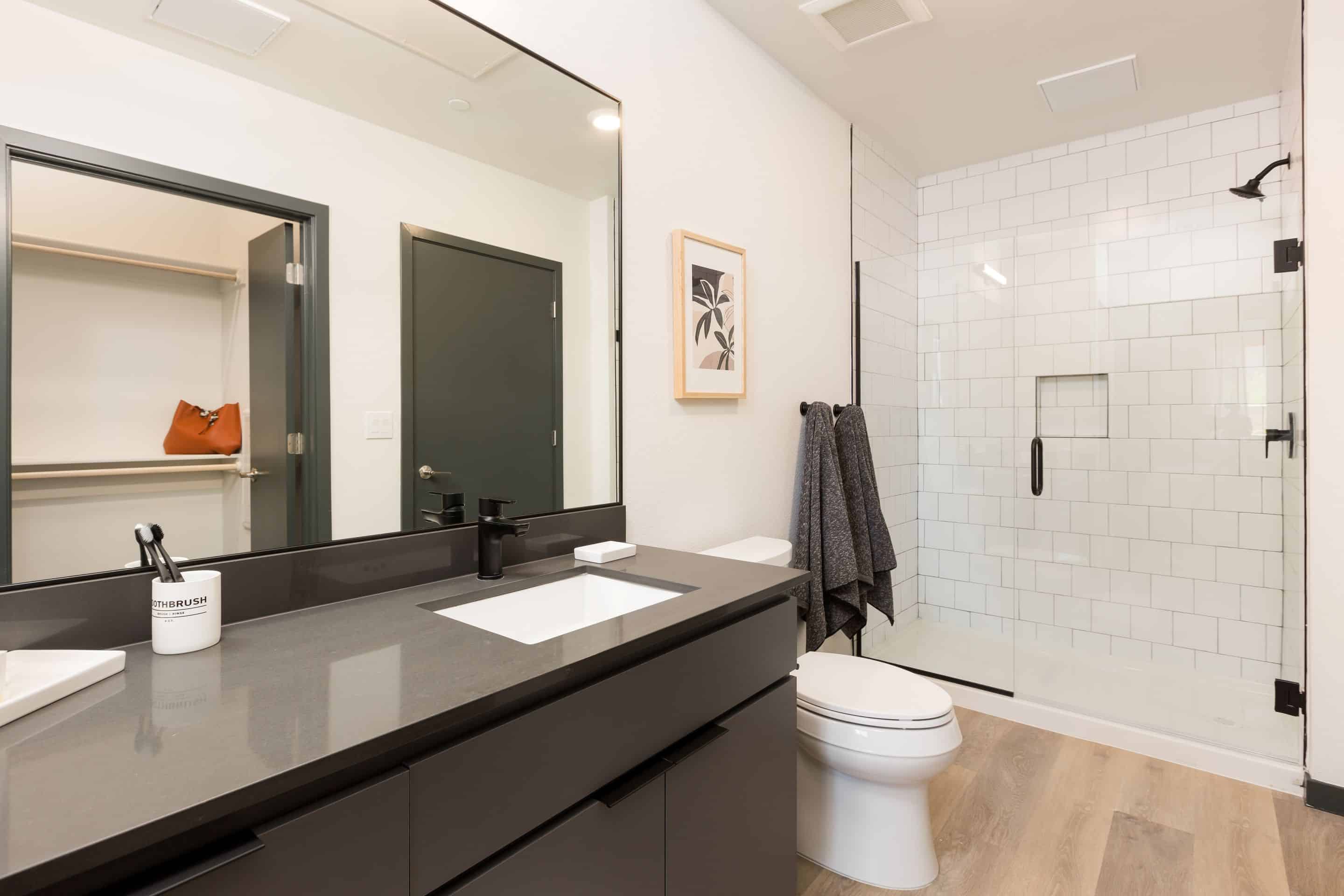 Modern bathroom in Home - Lake Cameron with a black vanity, rectangular mirror, walk-in shower featuring white subway tiles, toilet, and light wood flooring. Closet with a brown bag visible through an open door.