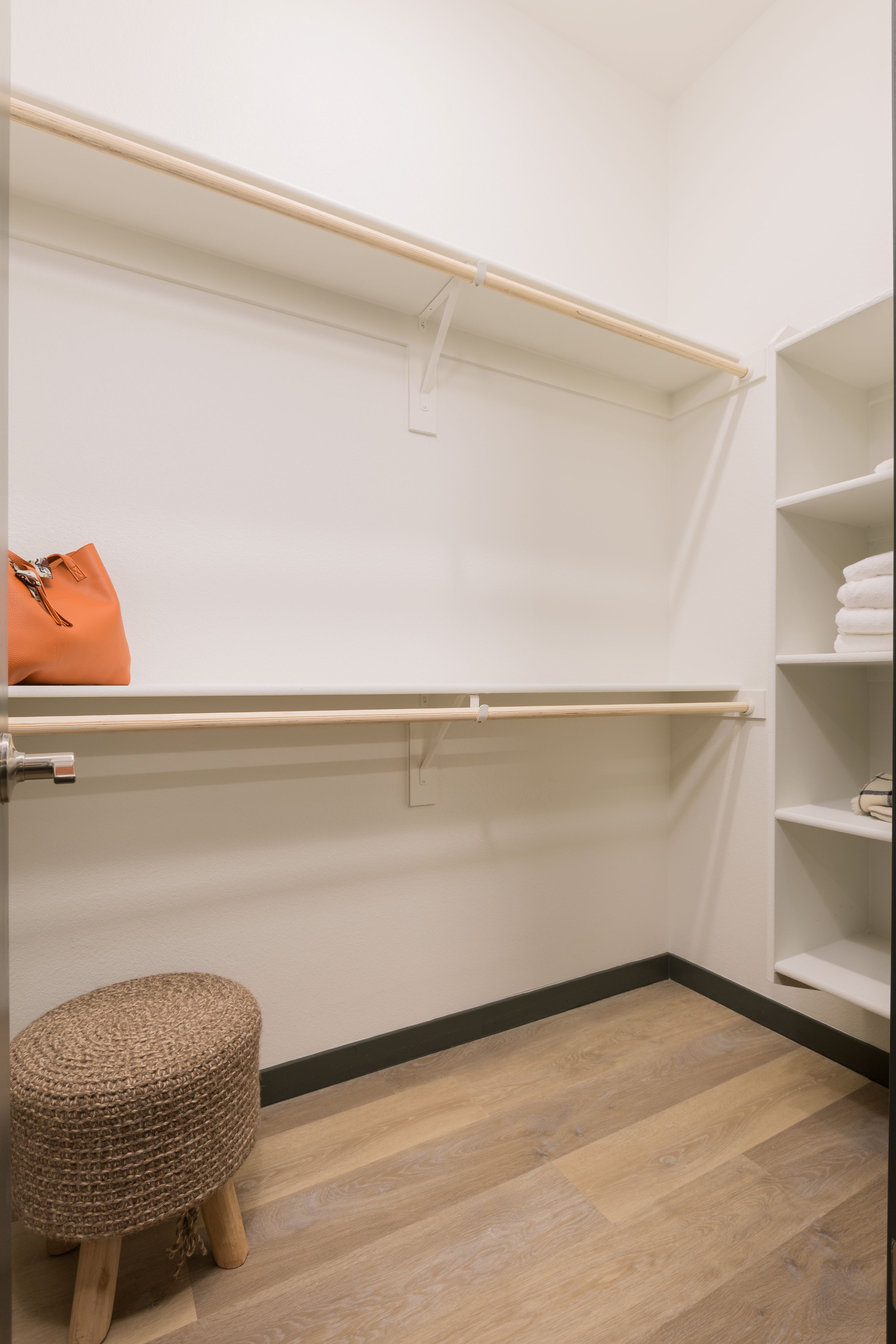 A walk-in closet in this Home - Lake Cameron features empty wooden shelves, a small woven stool, a single orange handbag, and folded towels on a white shelving unit.