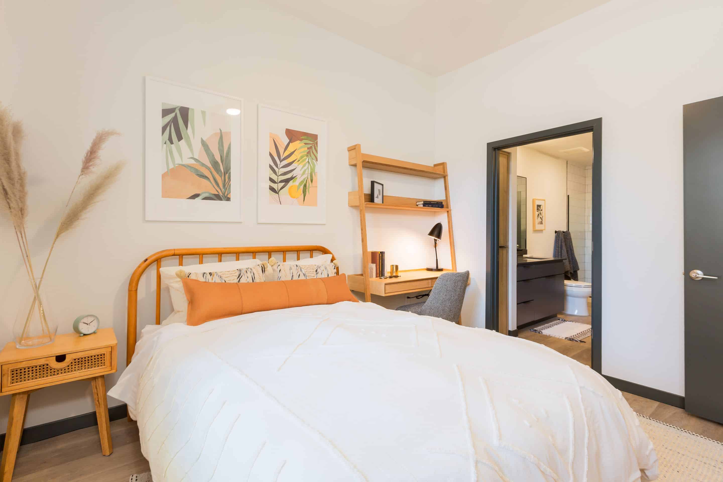 A modern bedroom at Home - Lake Cameron features a wooden bed, white bedding, a small nightstand, a wall-mounted desk, and an open door to the bathroom. Two framed botanical prints hang above the bed.