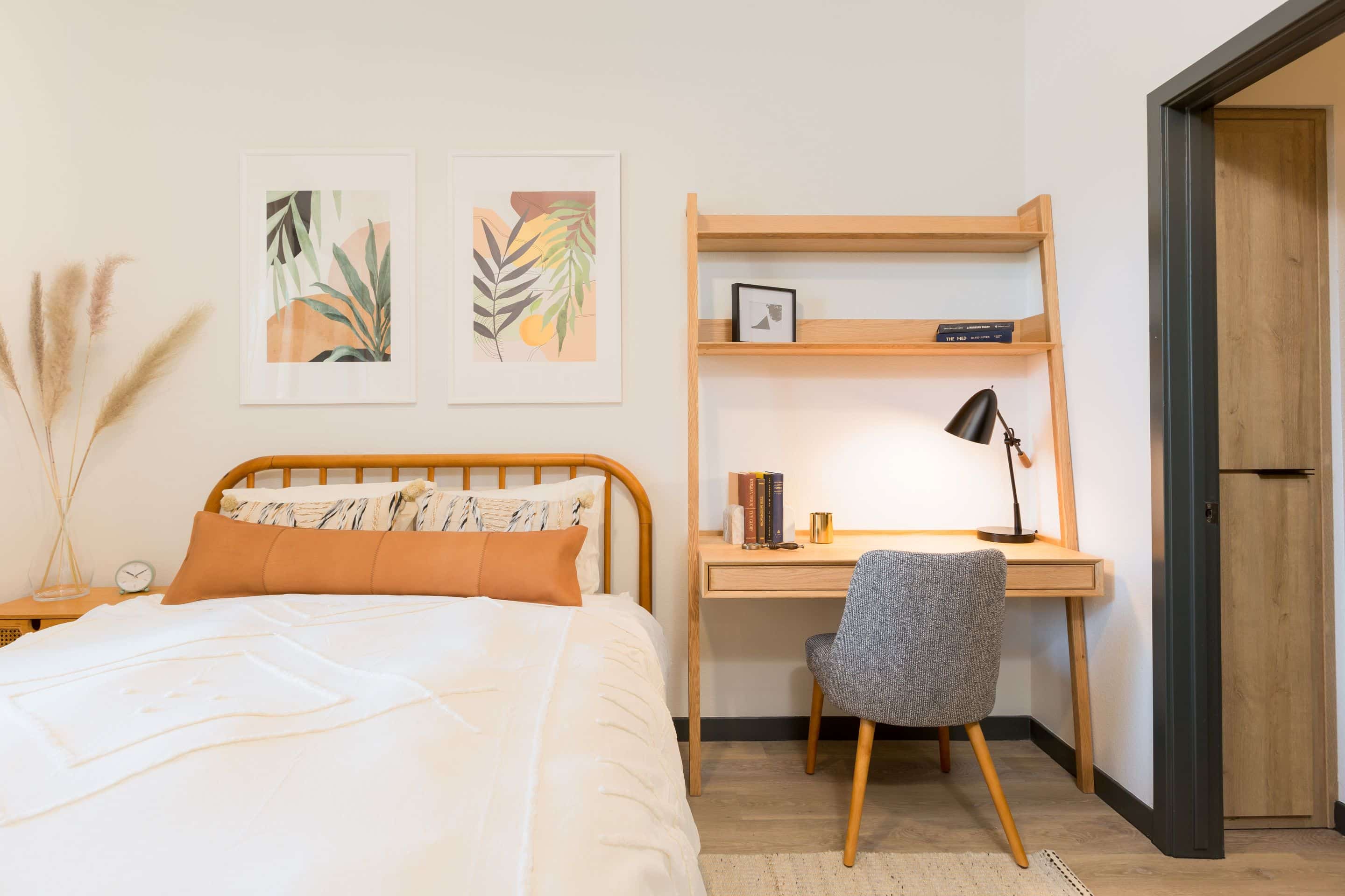 A neatly made bed with a wooden headboard is next to a wooden desk with a lamp, books, and a gray chair; wall art and pampas grass decorate this cozy Home at Lake Cameron.