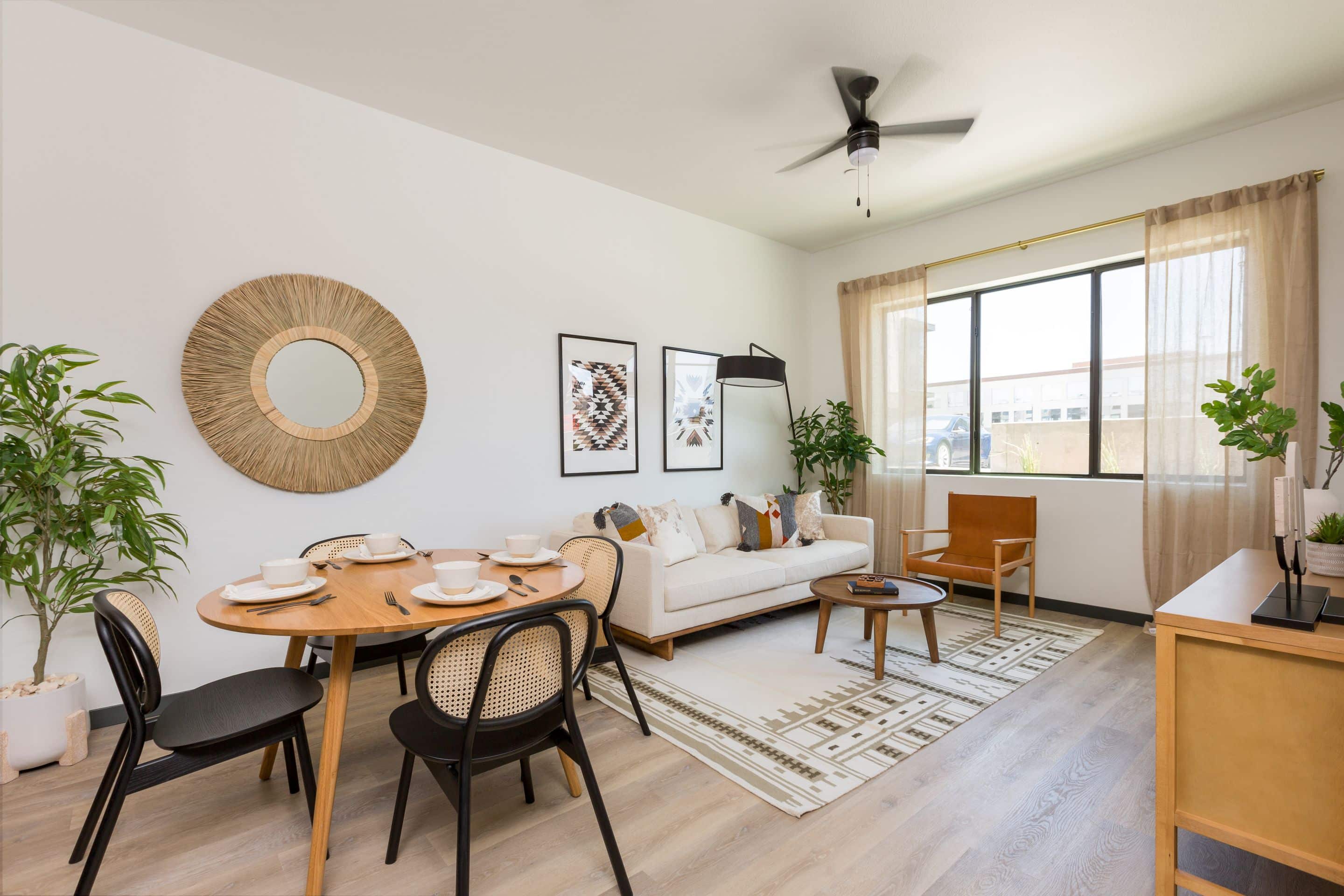 Bright living and dining area in Home - Lake Cameron with modern decor, a round dining table set for four, sofa, large window with sheer curtains, lush plants, and stylish wall art.