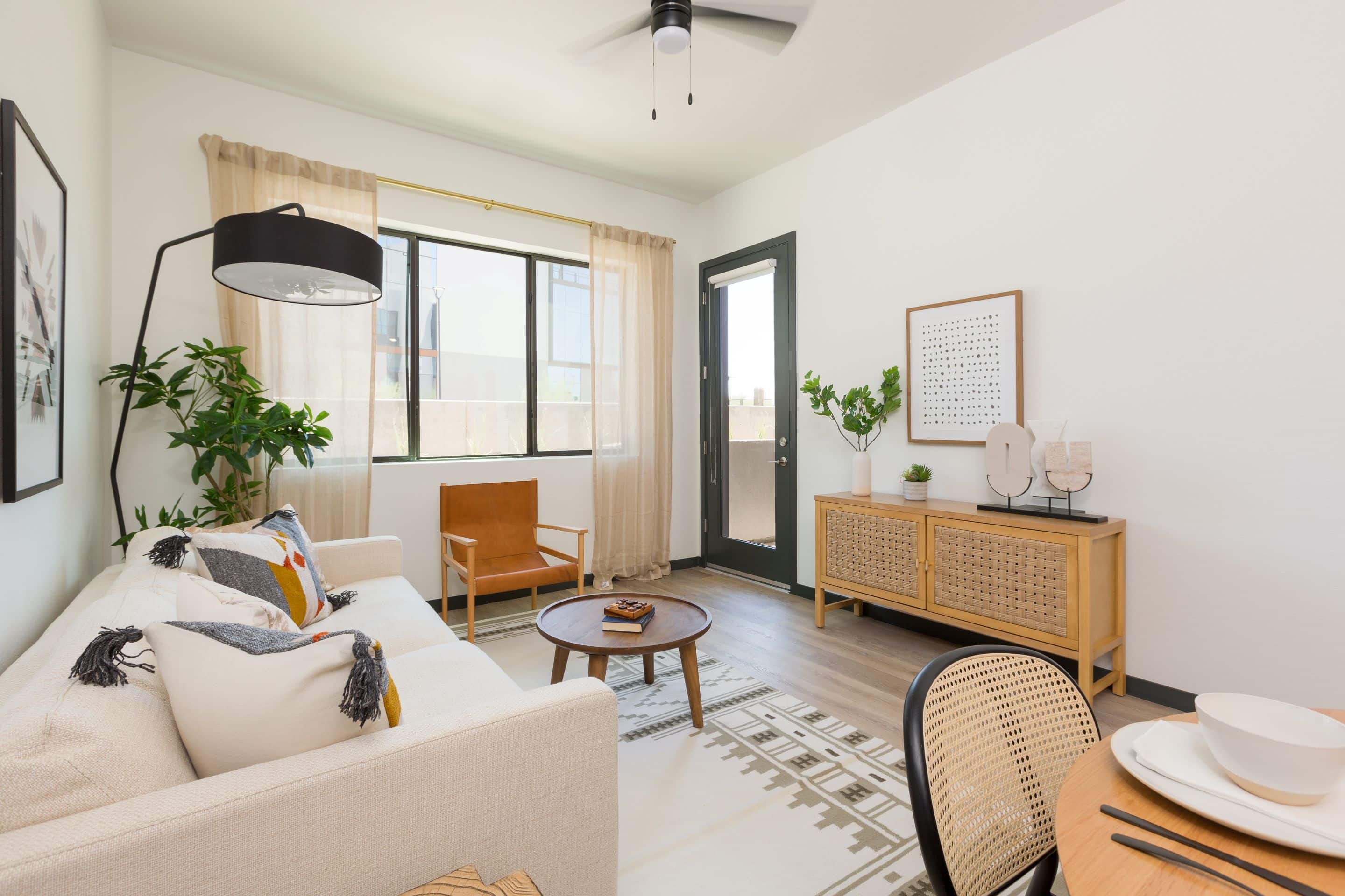 A modern living room at Home - Lake Cameron features a white sofa, wooden chair, coffee table, and credenza. Large windows with beige curtains let in natural light; neutral colors and plants decorate the inviting space.