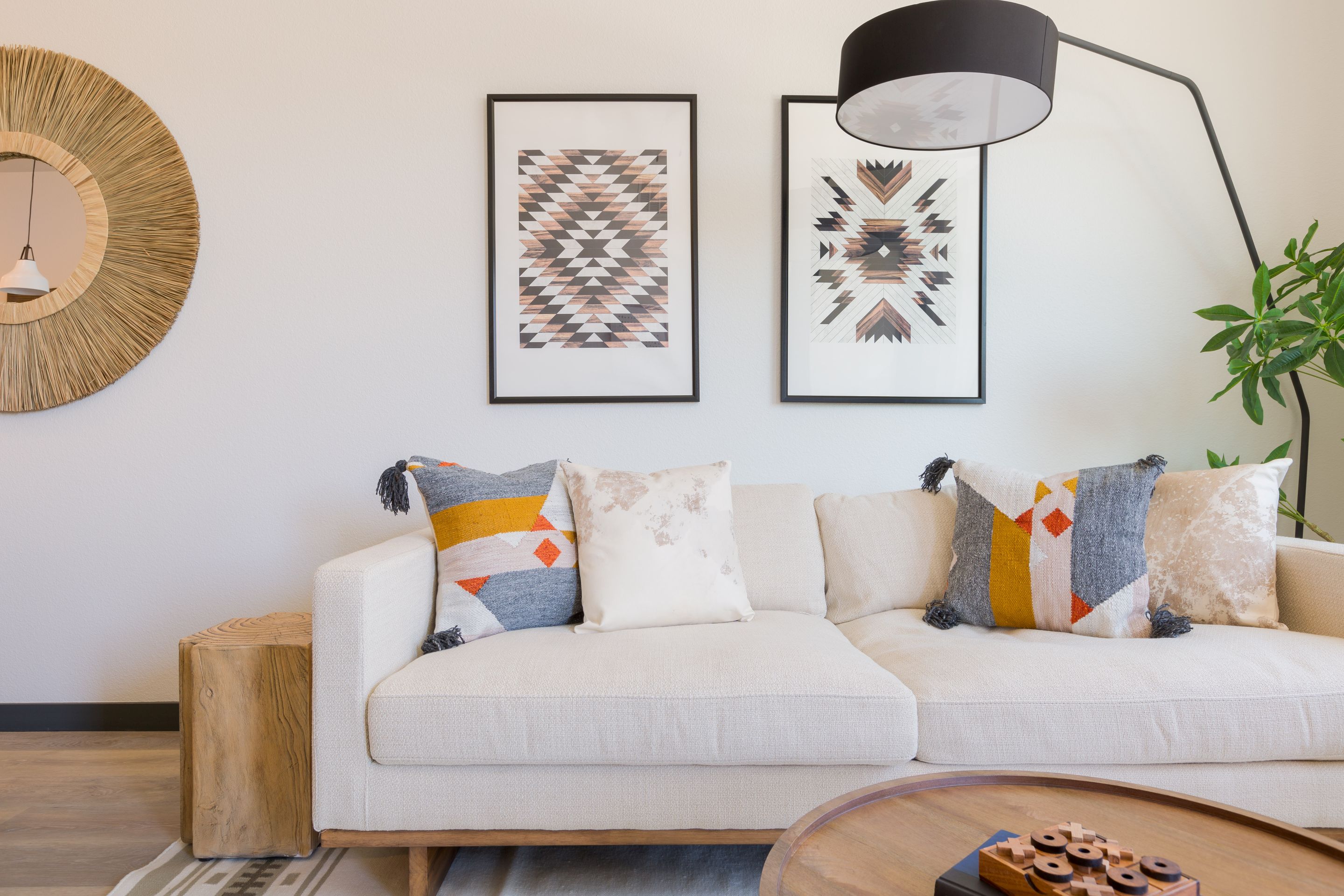 A beige sofa with patterned pillows sits against a wall with two geometric art pieces. A round mirror, wooden side table, lamp, and potted plant complete this modern Home at Lake Cameron.