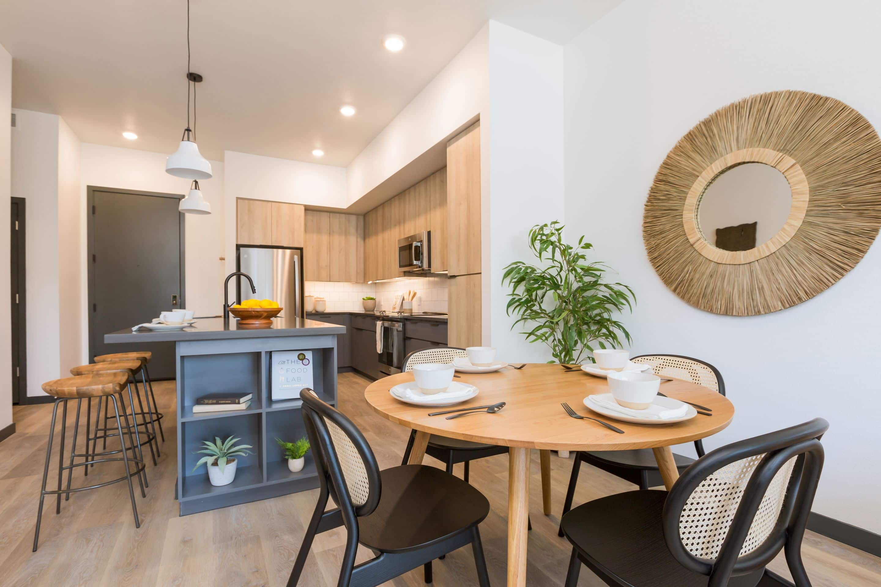 Modern kitchen and dining area in Home - Lake Cameron with wooden cabinets, a round table set for four, island with stools, potted plant, and a decorative round mirror on the wall.