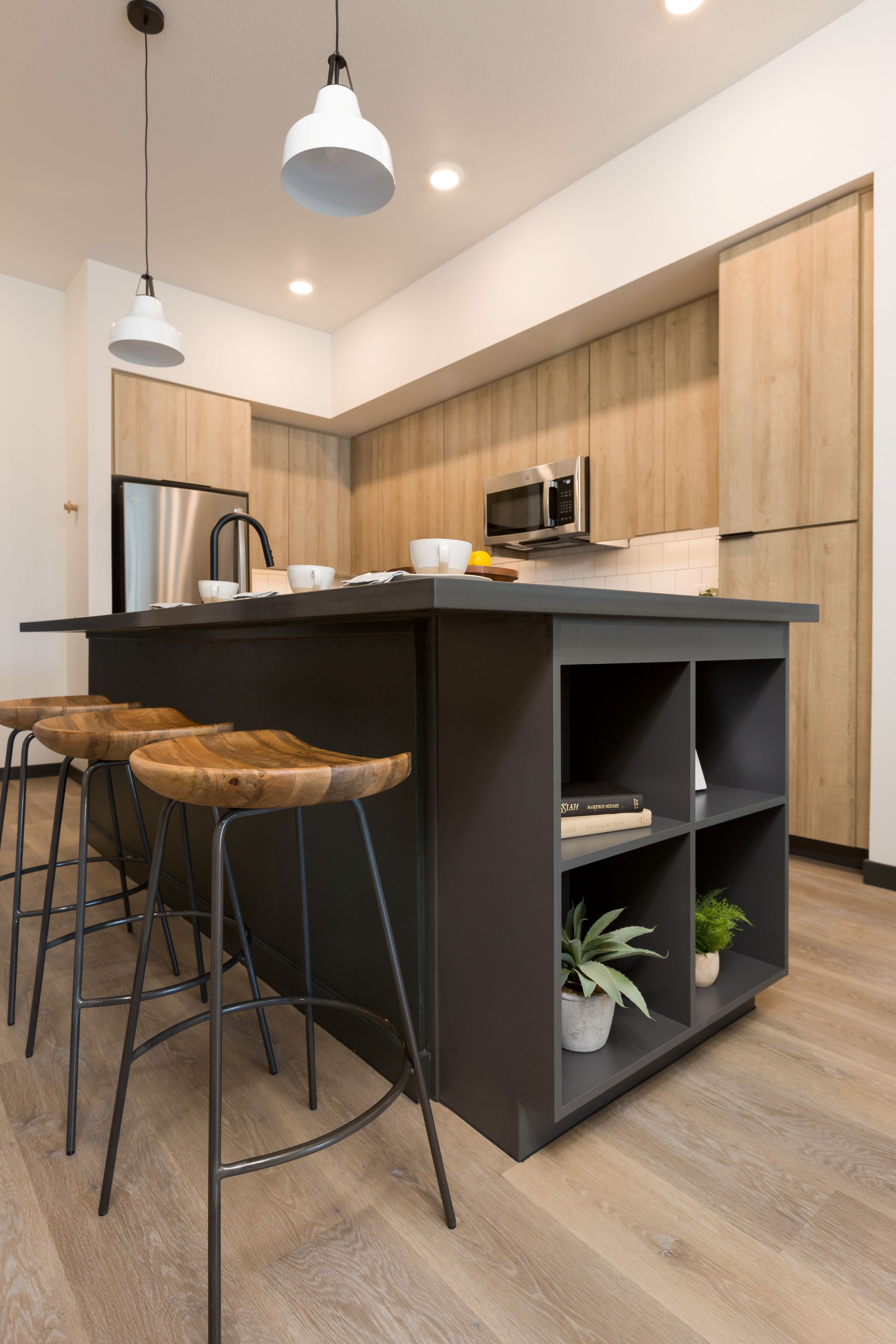 Modern kitchen with light wood cabinets, a black island with open shelving, barstools, pendant lights, and a few small plants and cups on the countertop—perfect for stylish living at Home - Lake Cameron.