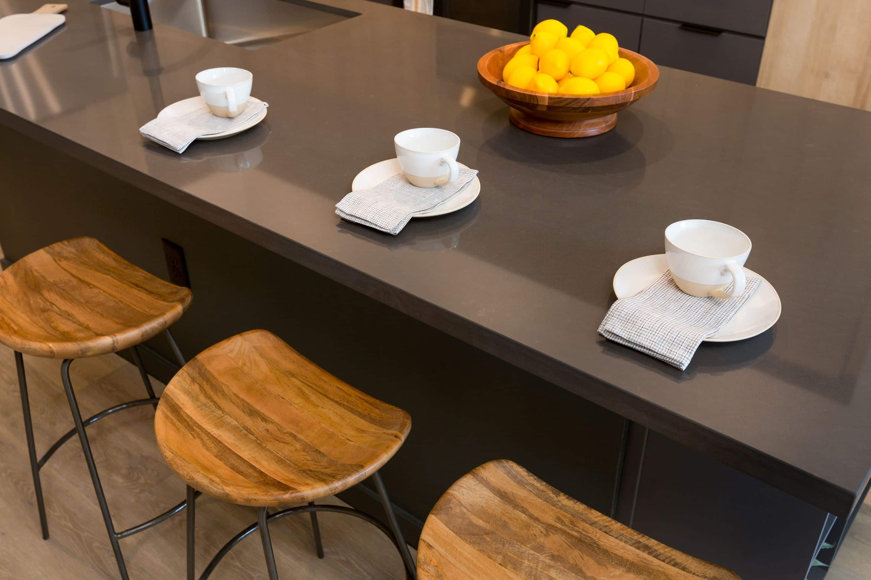 In this Home - Lake Cameron kitchen, three wooden stools line the island, topped with tea cups on saucers, napkins, and a wooden bowl brimming with bright lemons.