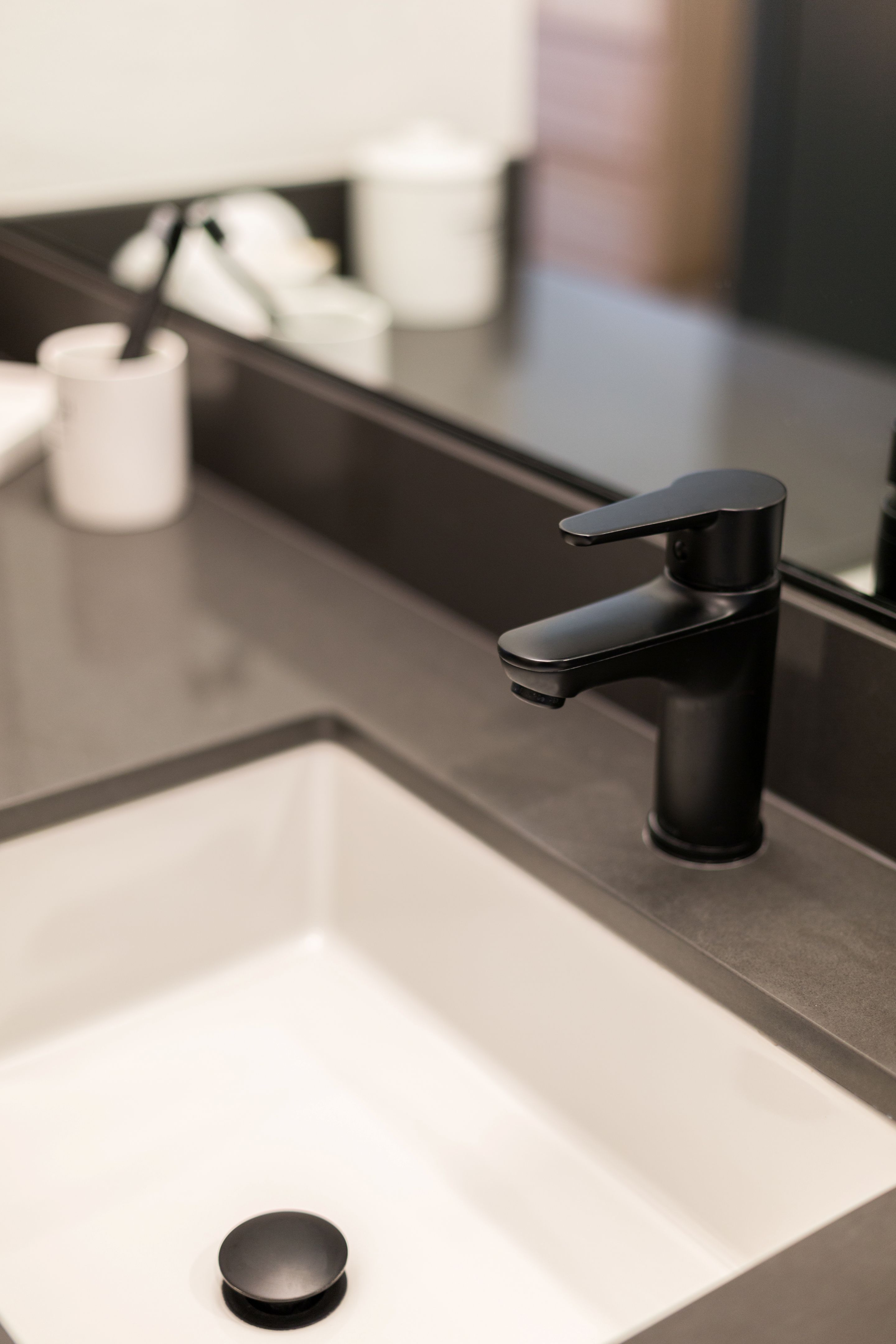 A modern black faucet and white rectangular sink on a dark countertop, with a toothbrush holder and other bathroom items reflected in the mirror, add sleek style to this Home - Lake Cameron retreat.
