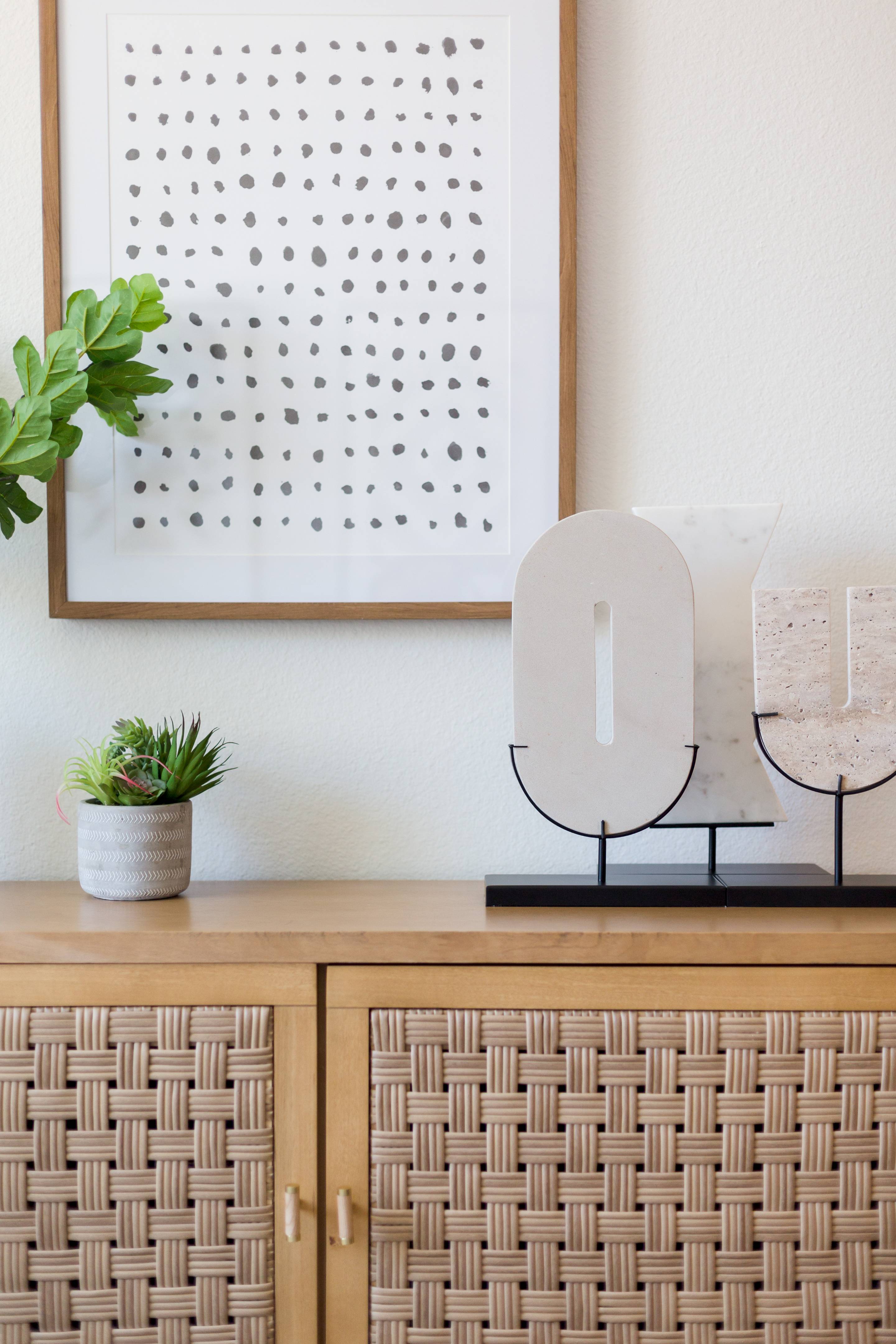 A light wood cabinet with a woven pattern holds a small potted plant and abstract decorative objects, adding modern charm to your Home - Lake Cameron. Above it hangs framed art featuring a grid of black dots.