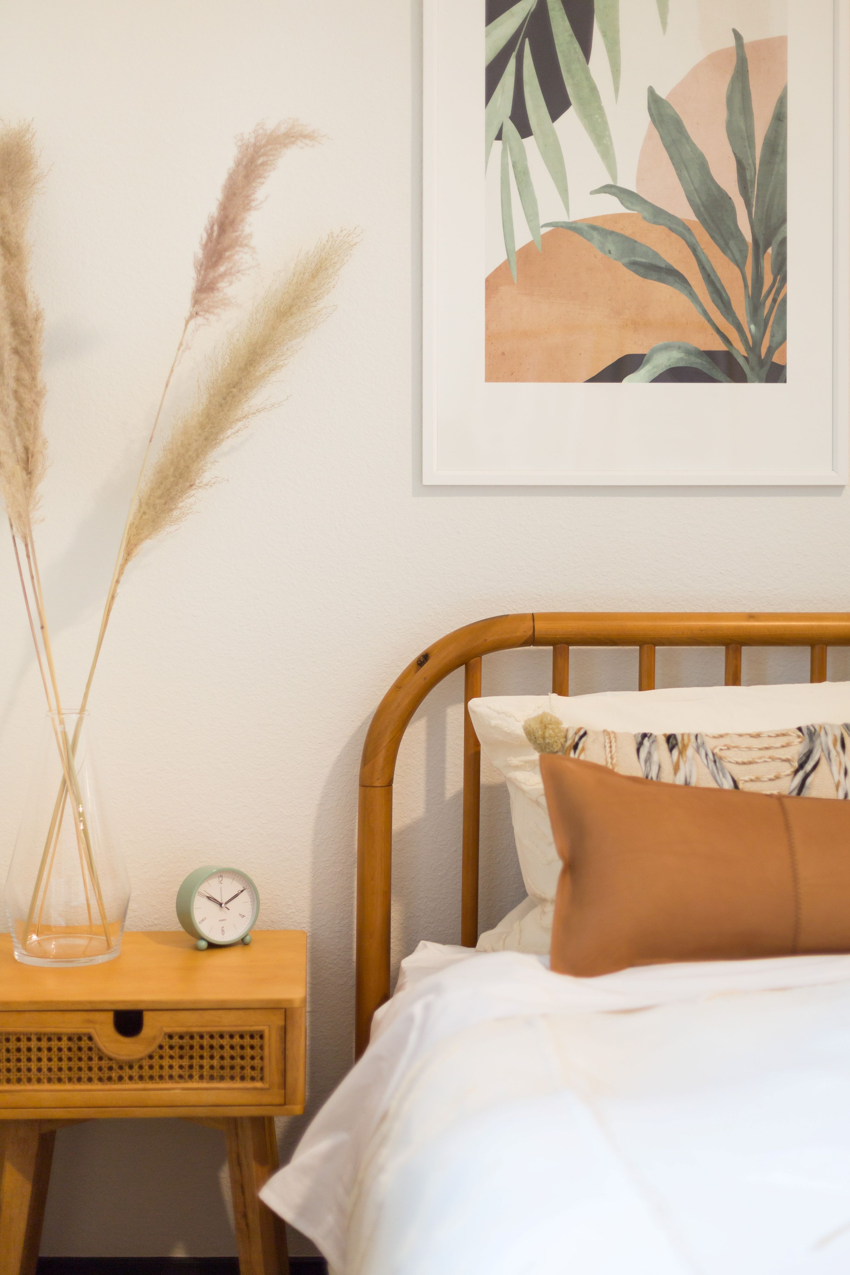A neatly made bed with white linens and brown pillows sits beside a wooden nightstand holding a clock, dried grasses in a vase, and a framed botanical print—inviting comfort and style to this Home near Lake Cameron.