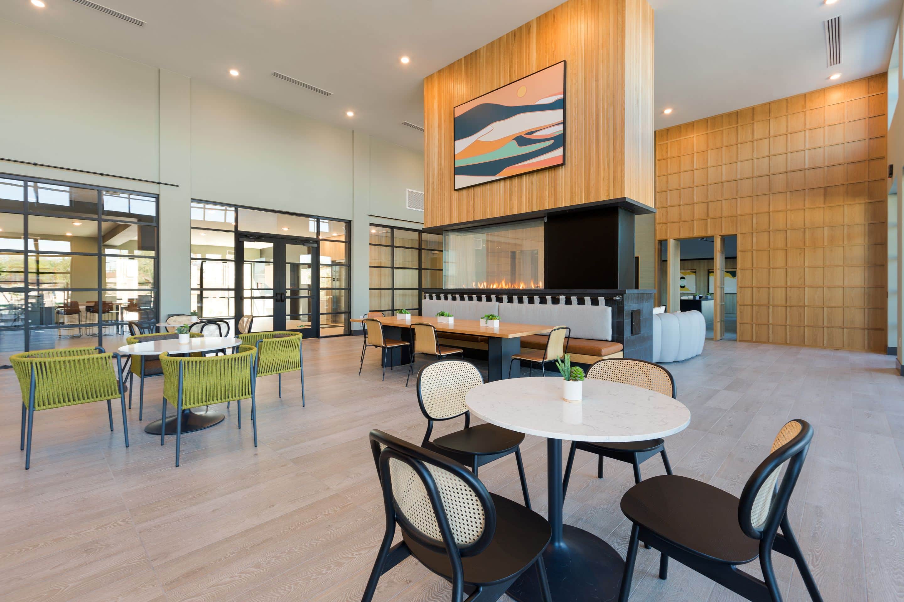 Modern communal lounge at Home - Lake Cameron with round tables, green and black chairs, light wood flooring, a large fireplace, and abstract art on a wood-paneled wall. Expansive windows fill the space with natural light.