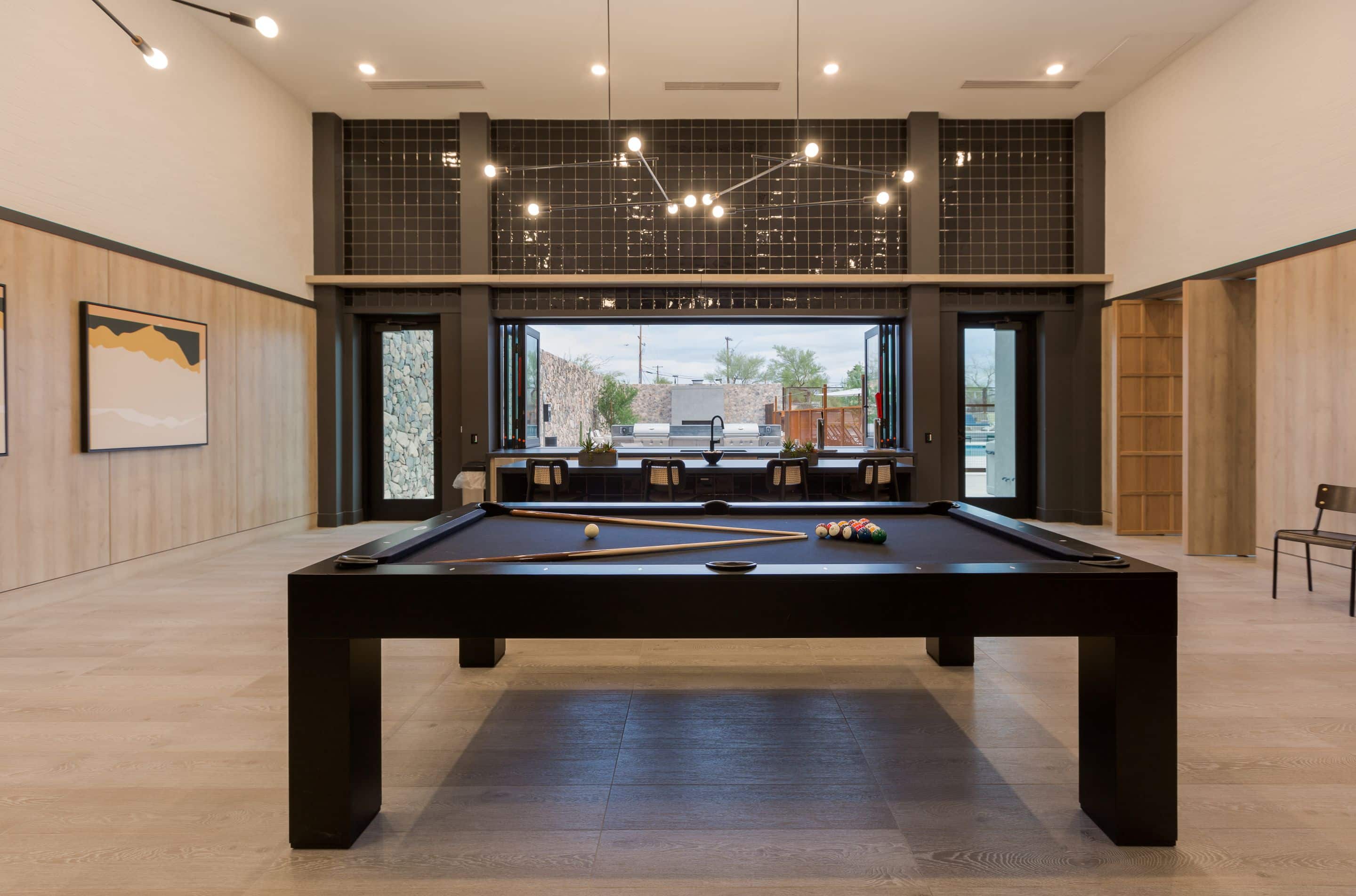 A modern game room at Home - Lake Cameron features a sleek black pool table in the center, cues on the table, minimalist decor, open folding doors, and a stunning view of the outdoor patio area.