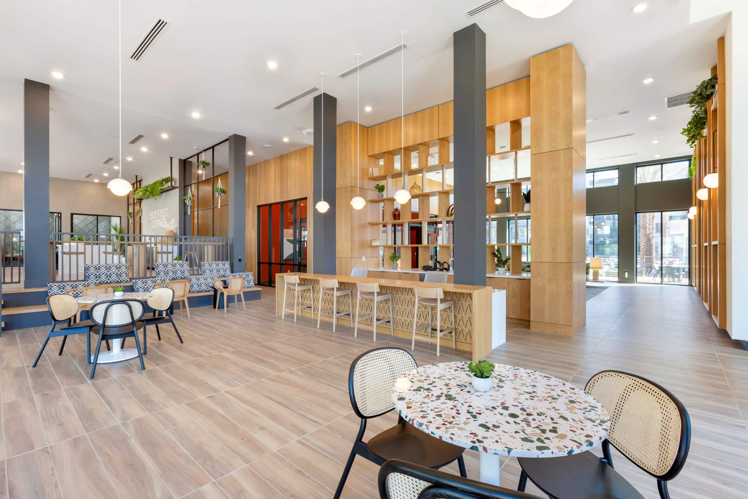 Modern communal lounge area at Home - Lake Cameron with wooden flooring, tables and chairs, pendant lighting, and large windows for natural light. Shelving and cozy seating nooks line the back wall.