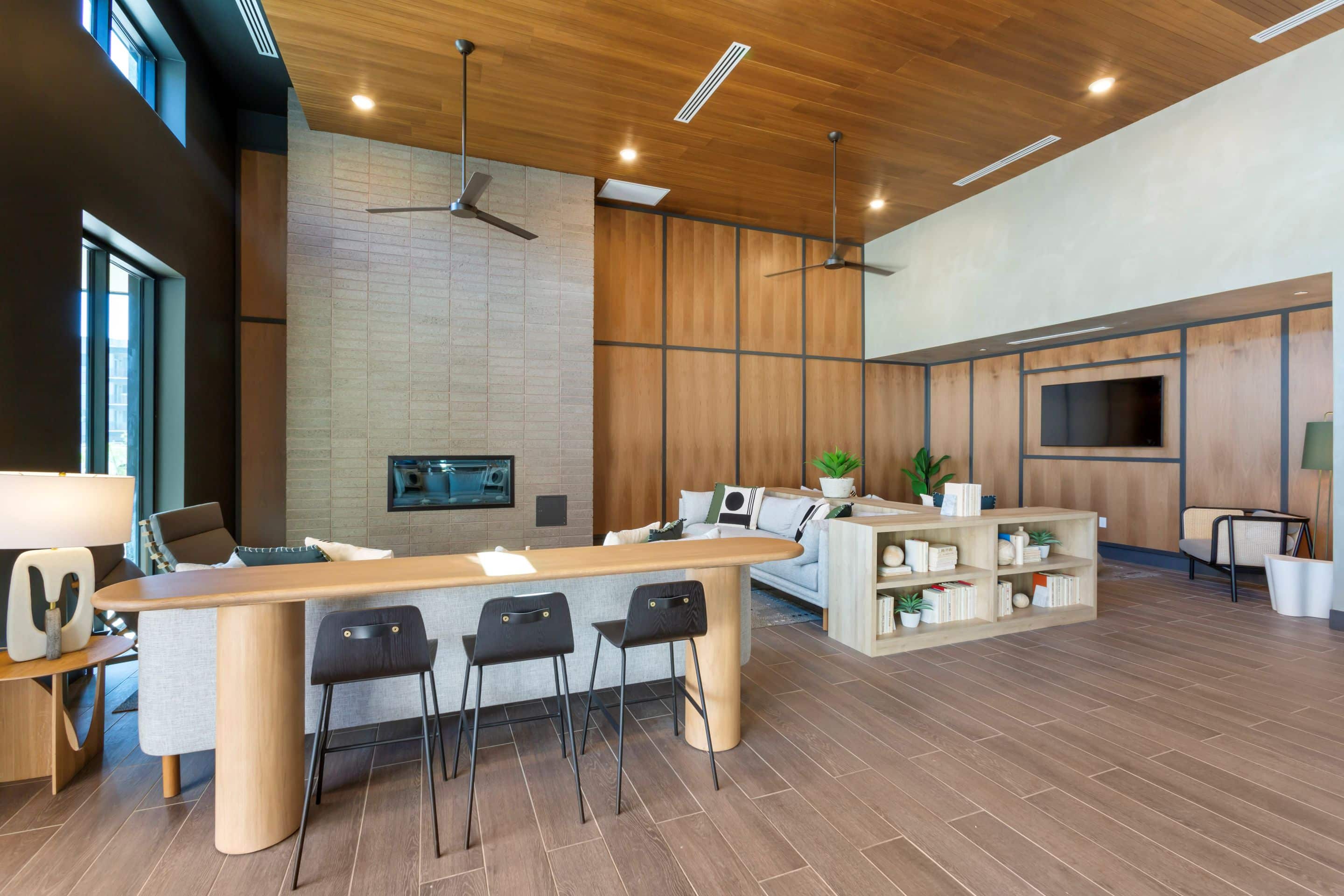 Modern living room in Home - Lake Cameron featuring a wooden ceiling, tile floors, fireplace, sofa, TV, and a light wood island with three black barstools. Built-in shelves on the island display books and decor.