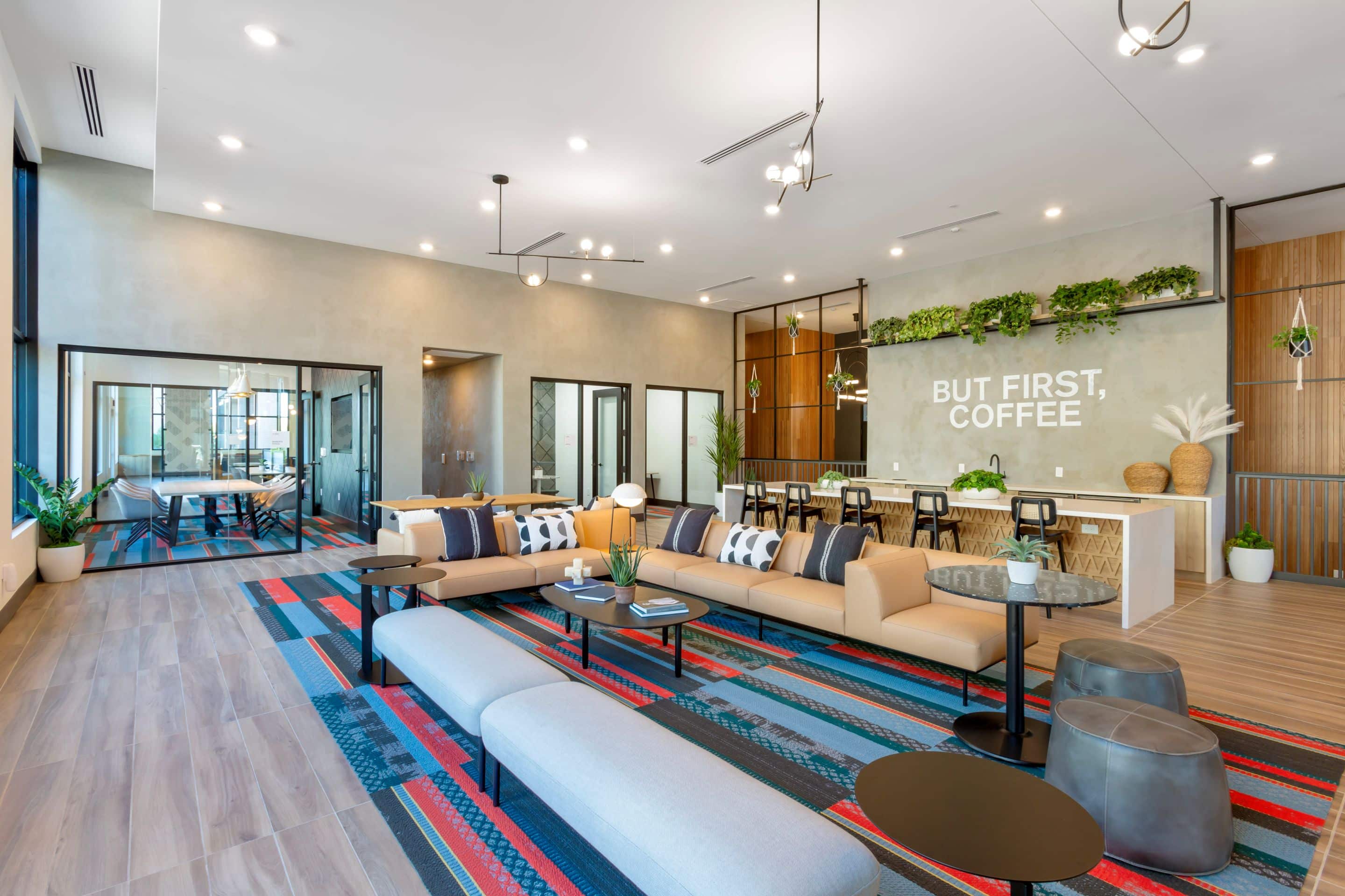 Modern lounge area with beige sofas, small tables, a colorful rug, and a coffee bar. A neon sign reads “But First, Coffee.” Large windows fill this Home - Lake Cameron space with natural light.