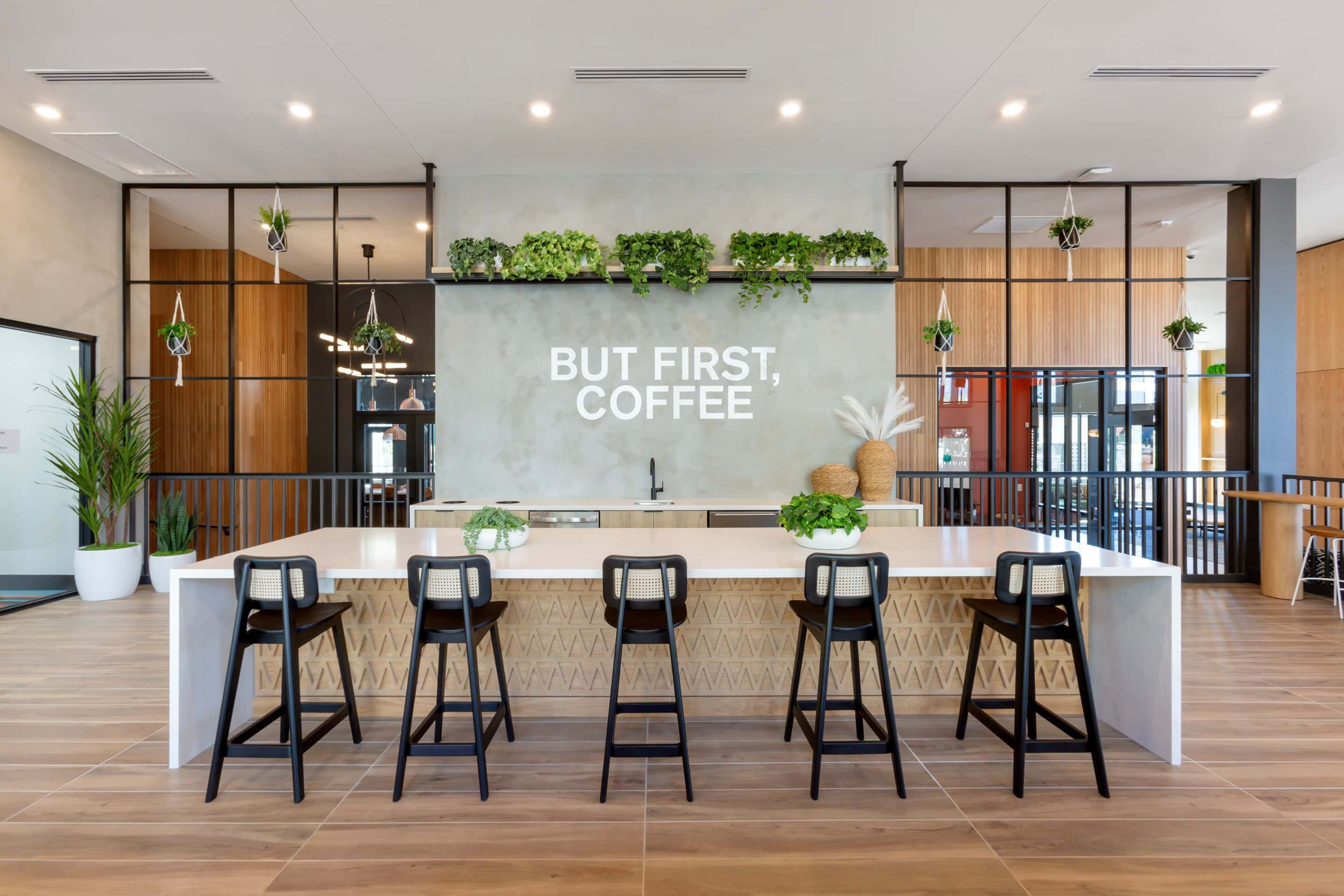 Modern coffee bar with a white counter, black chairs, potted plants, and a sign on the wall that reads "BUT FIRST, COFFEE." Enjoy this cozy spot at your Home - Lake Cameron.