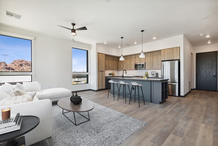Modern open-concept living room and kitchen at Scottsdale Entrada with wood floors, gray cabinets, a large sofa, bar stools, and natural light from two large windows.