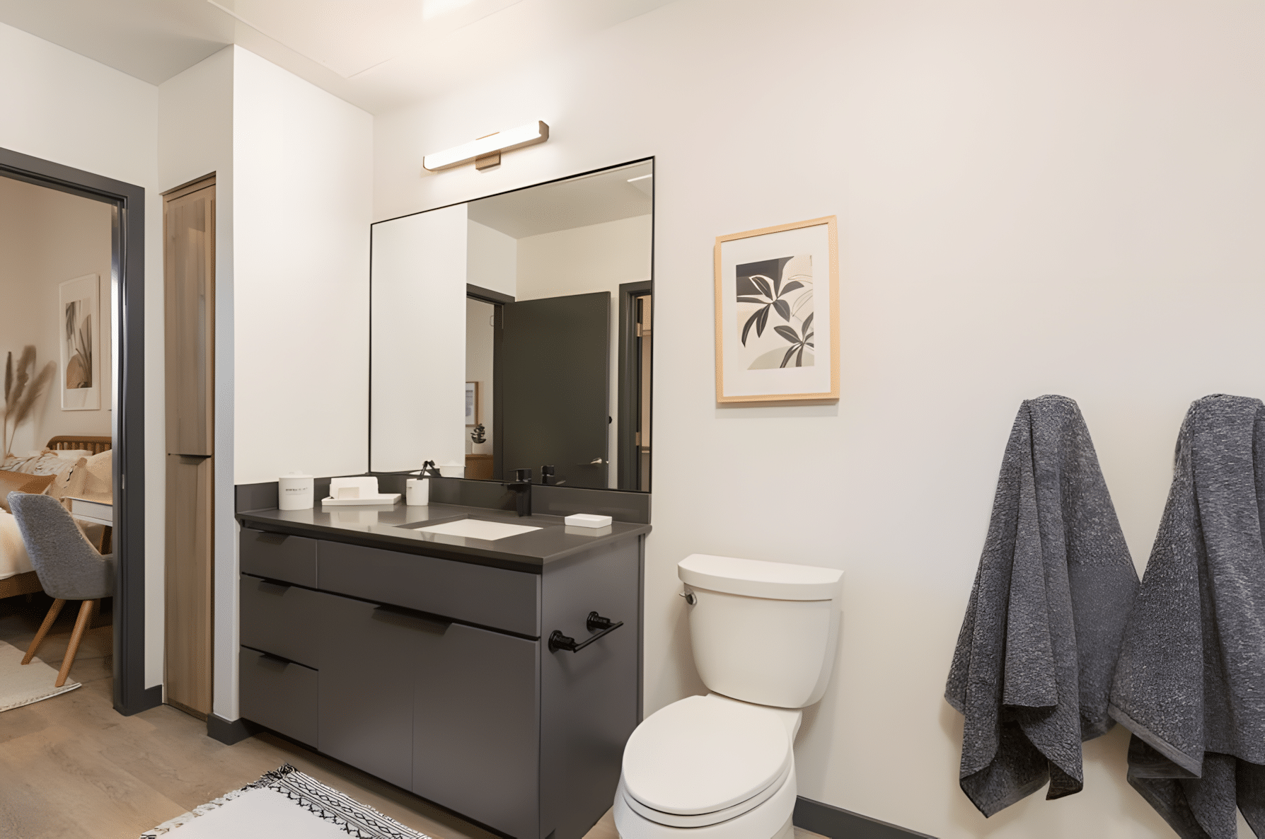 Modern bathroom with a dark vanity, large mirror, wall art, two gray towels on hooks, and a white toilet; bedroom visible through an open doorway.