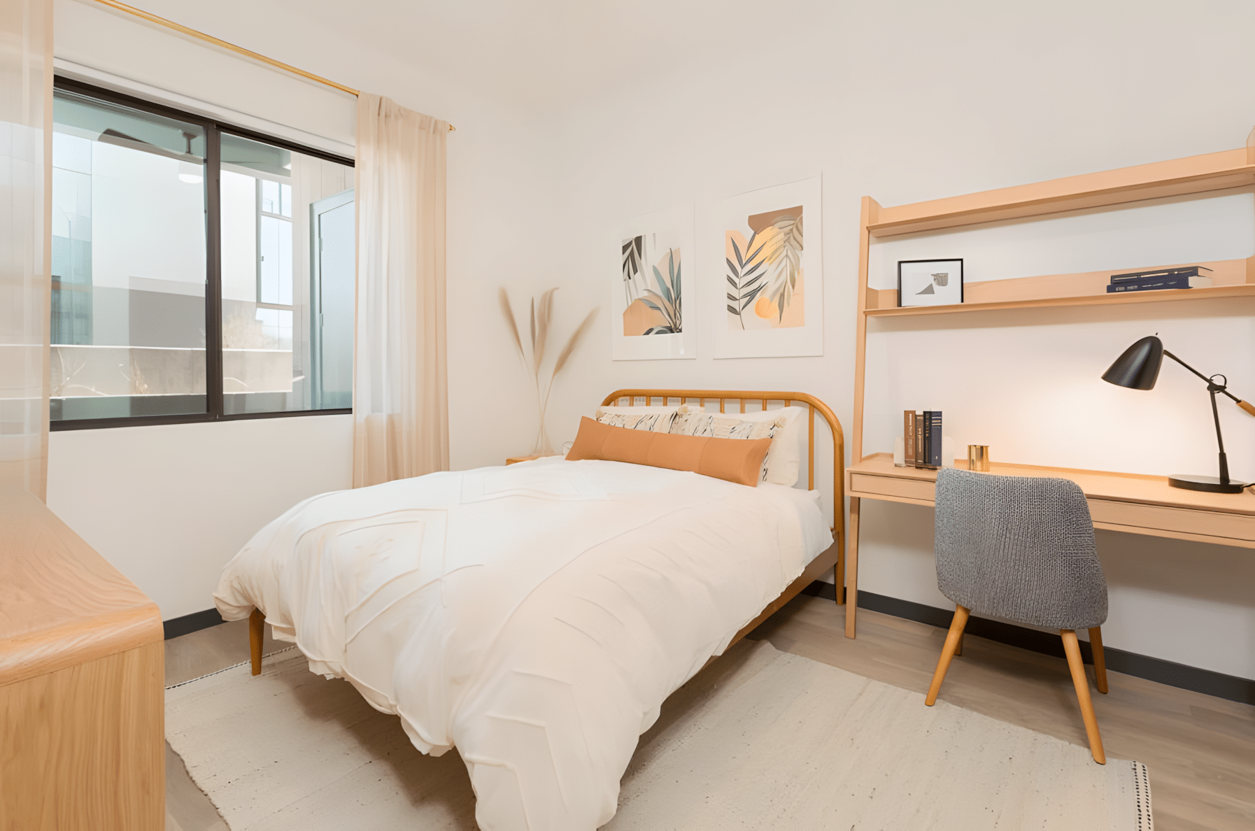 A neat bedroom with a bed, white bedding, a wooden desk and chair, wall art, open window, and natural light.