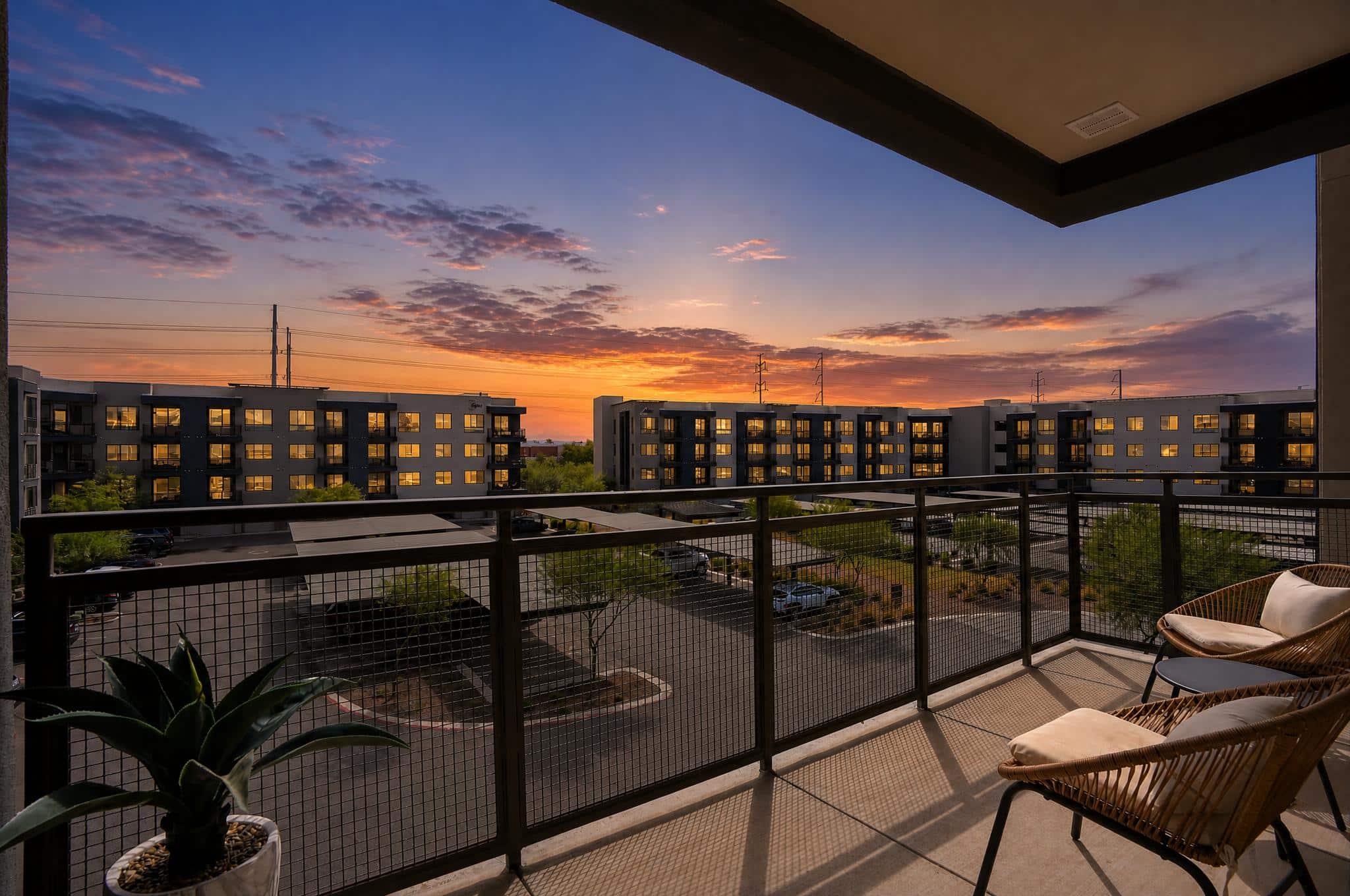 Apartment balcony with two chairs and a potted plant overlooks a parking lot and modern buildings at sunset, with a colorful sky in the background.