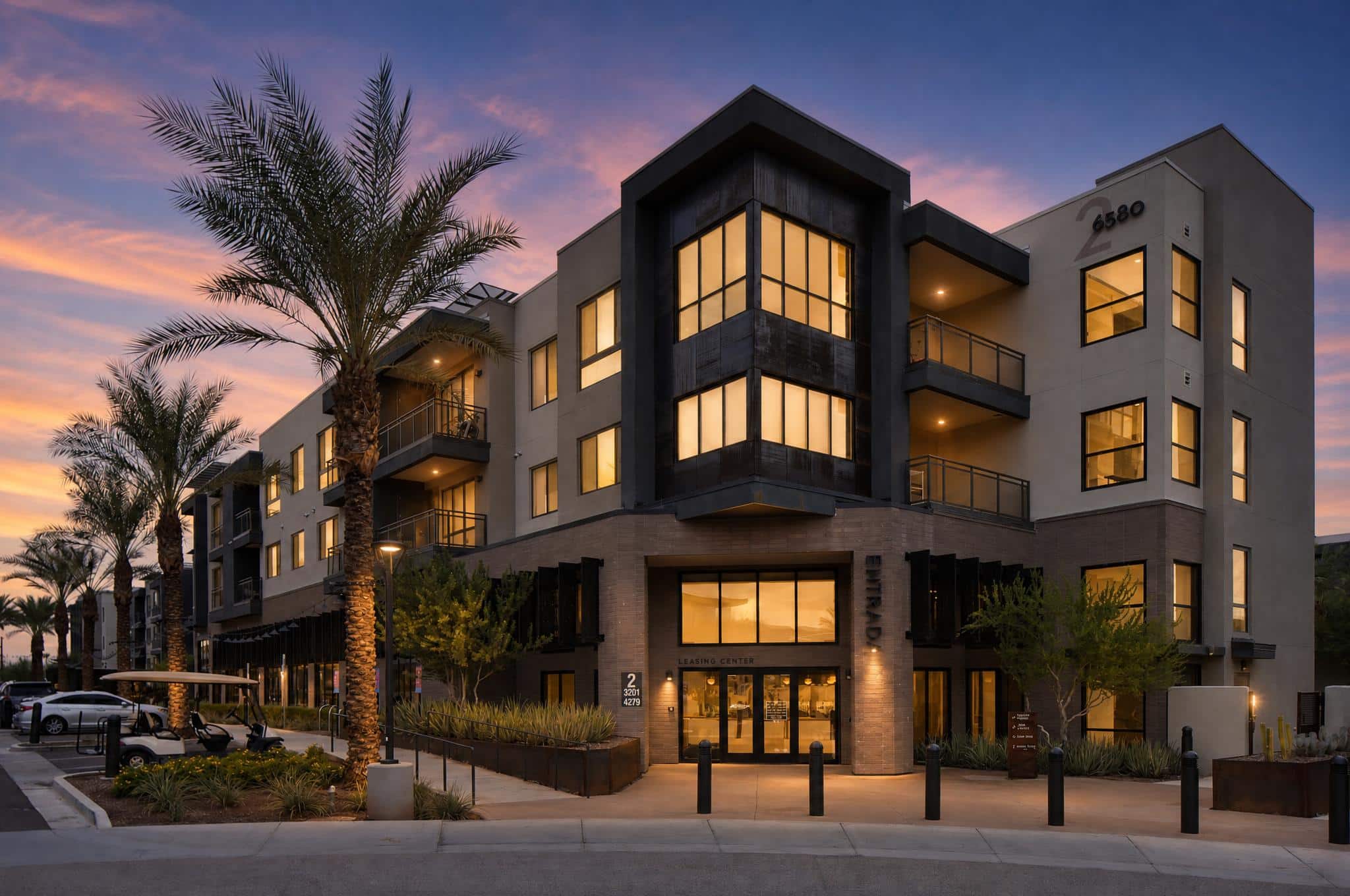 Modern three-story apartment building at dusk, with large windows lit from inside, palm trees, balconies, and main entrance visible.