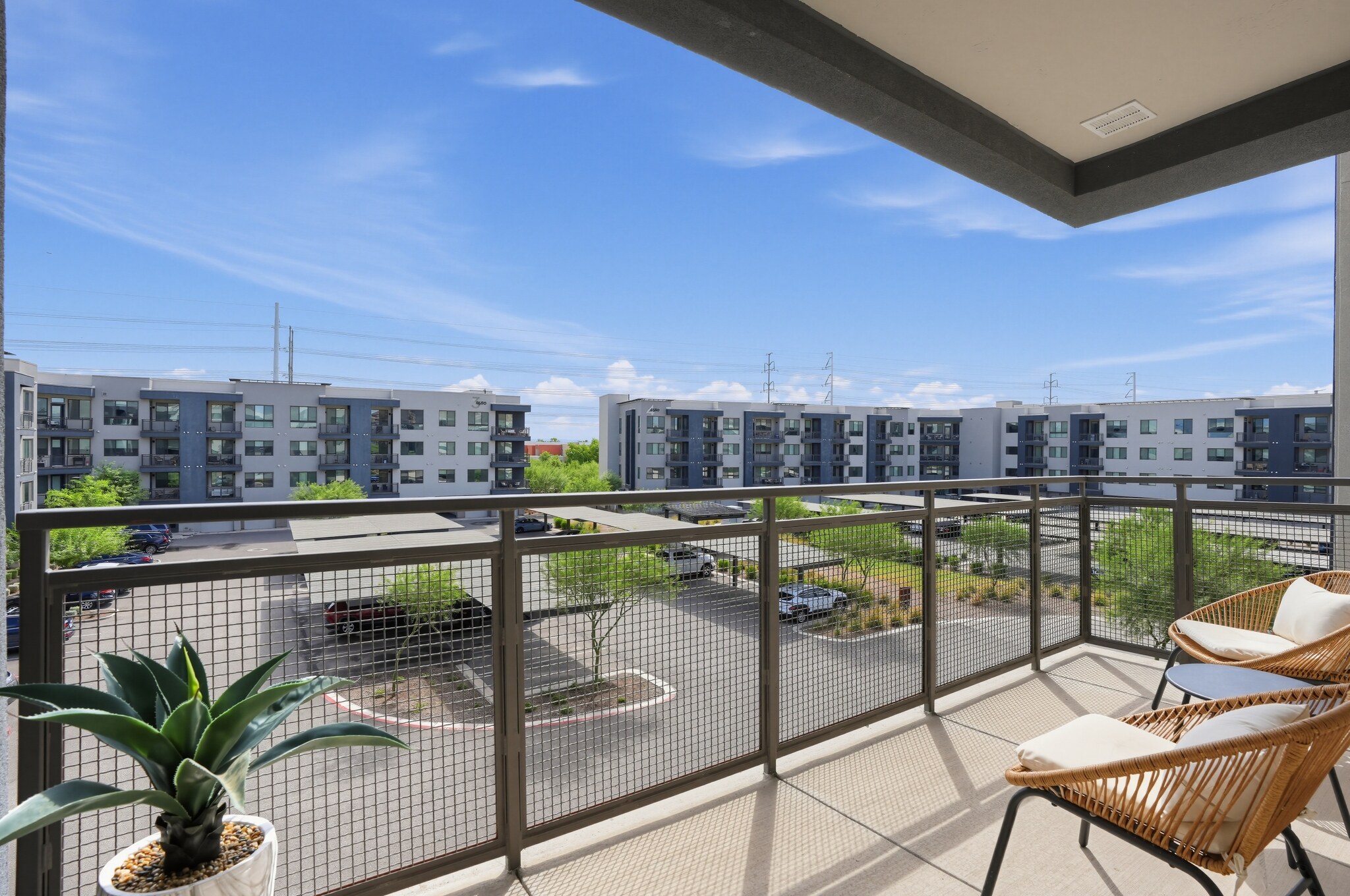 Modern apartment balcony with two chairs and a potted plant, overlooking a parking lot and adjacent apartment buildings under a clear blue sky.