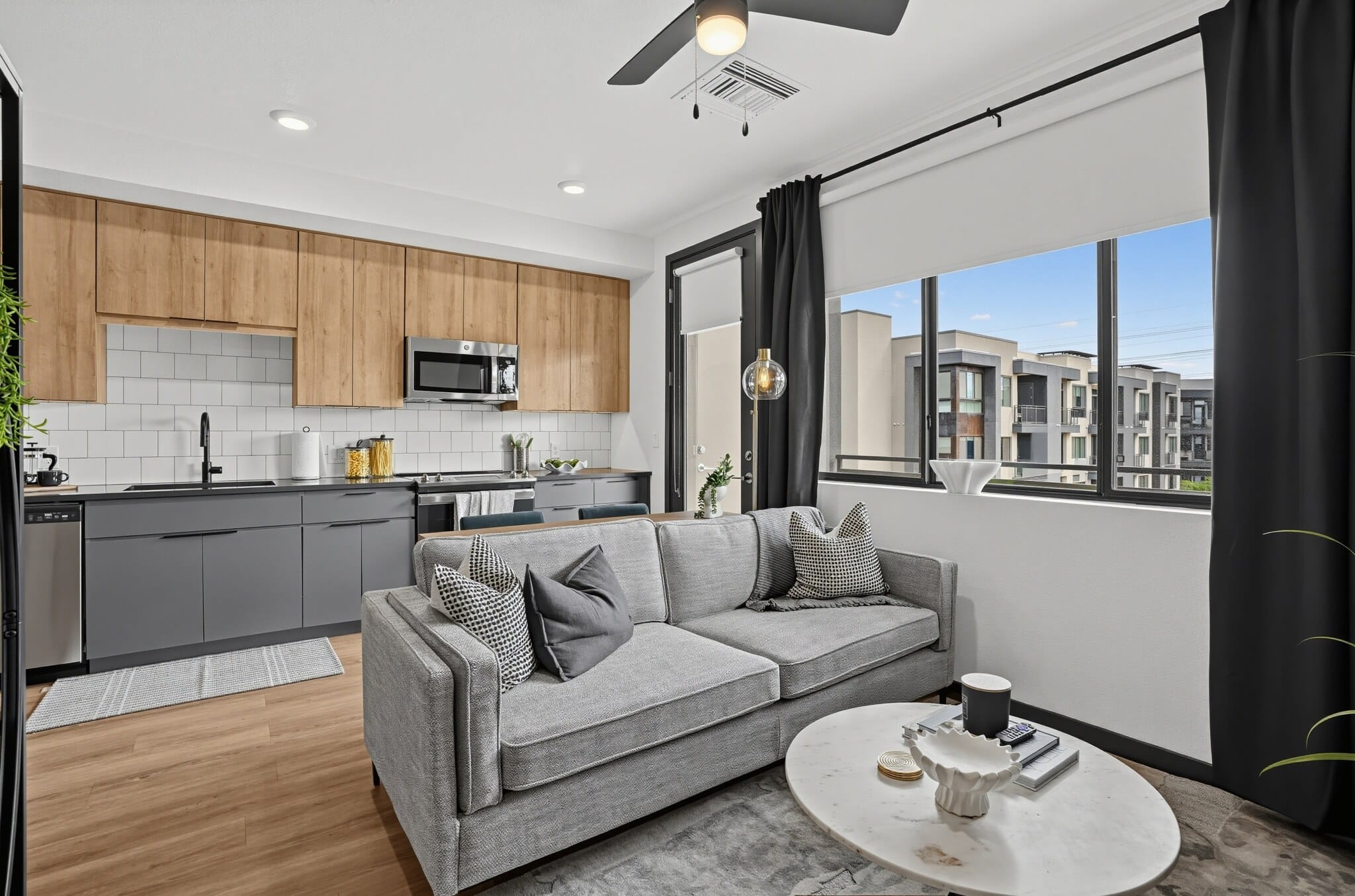 Modern apartment living room with a gray sofa, marble coffee table, and kitchen area with wood cabinets, black appliances, and large windows showing adjacent buildings.